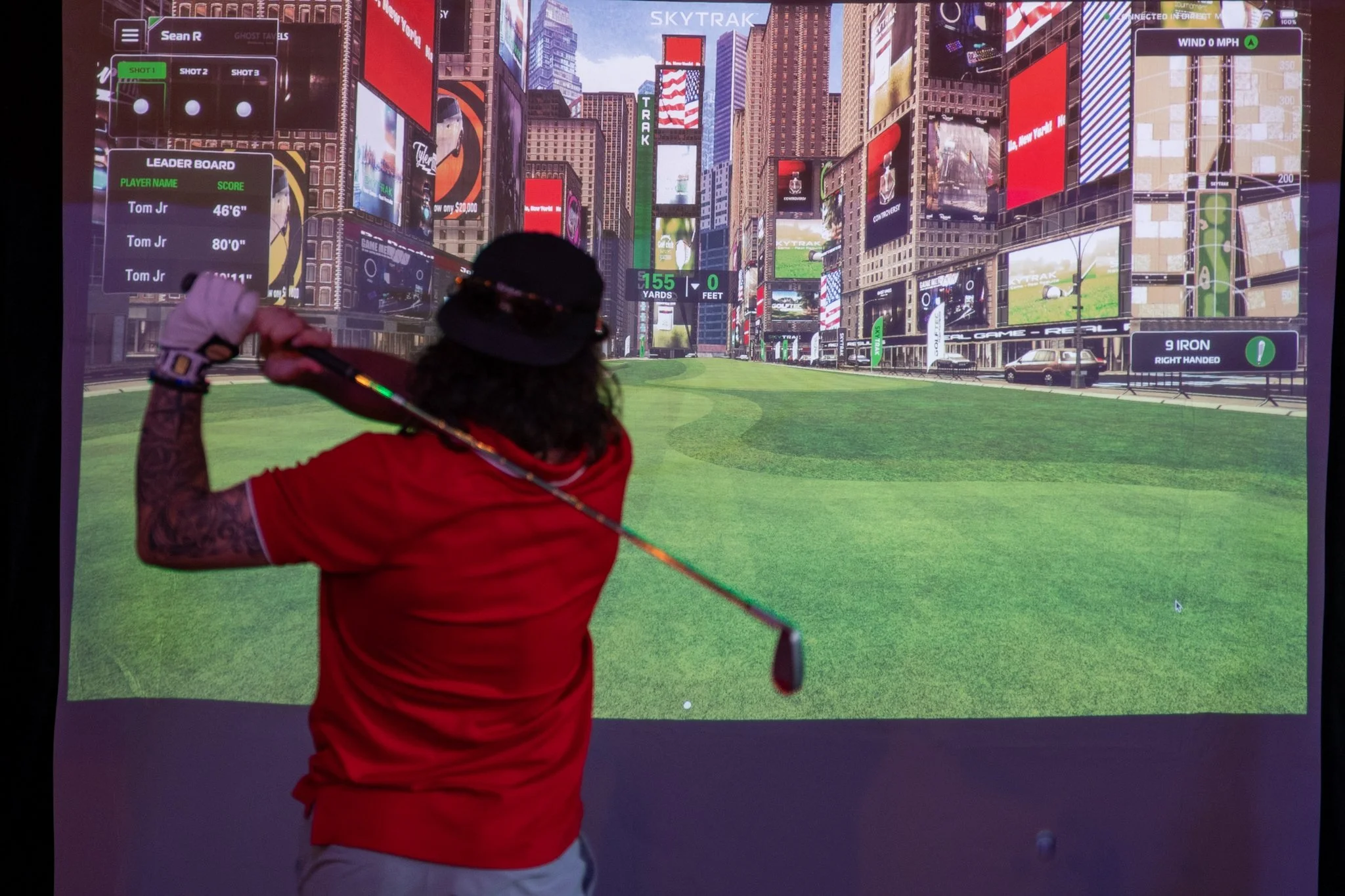 A person practicing golf using a simulator with a cityscape background of tall buildings and digital screens.