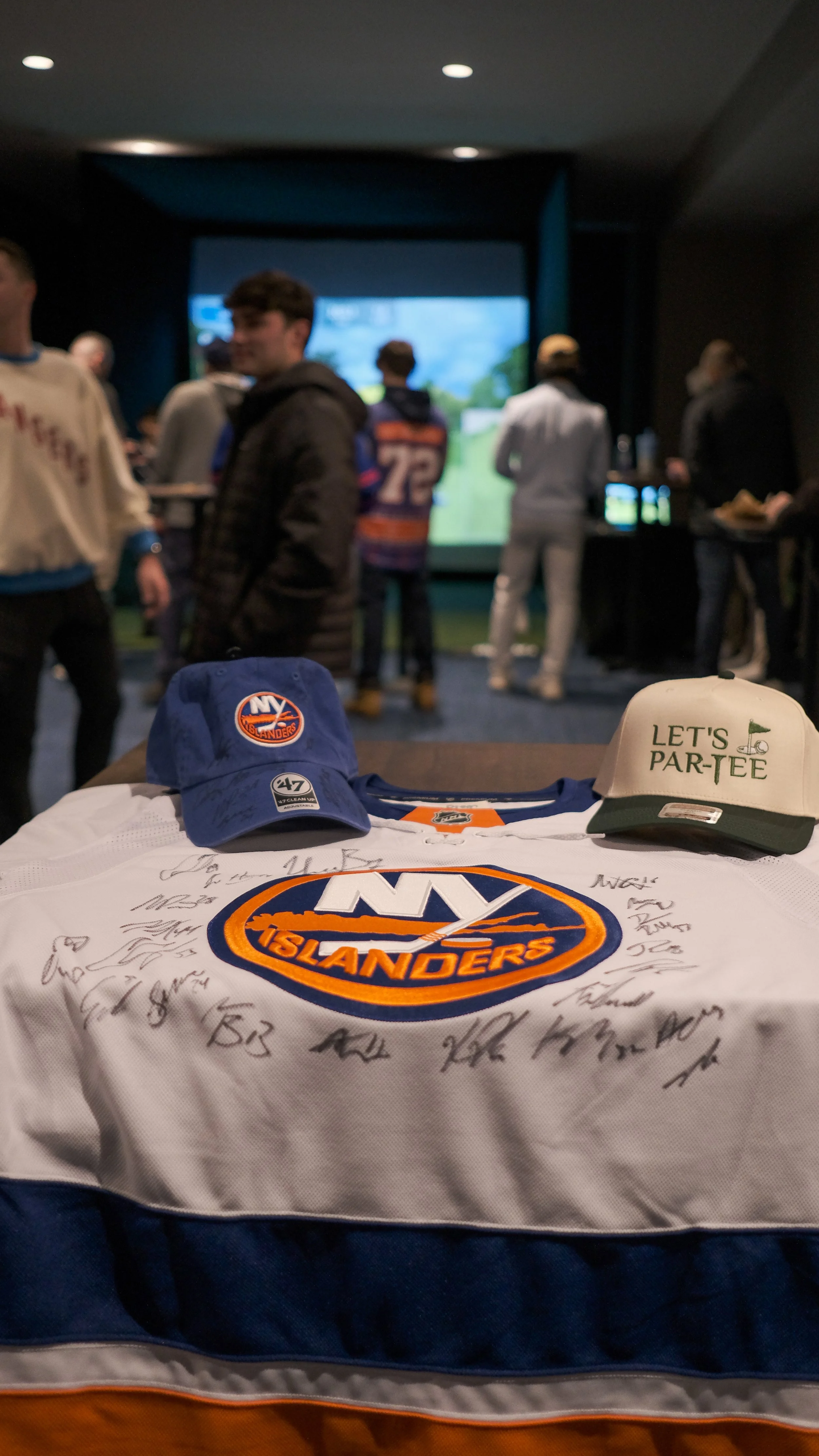 New York Islanders hockey jersey autographed, with two caps on top, one blue with Islanders logo and one white with 'Let's Par-Tee' text, displayed on a table at a gathering or event.
