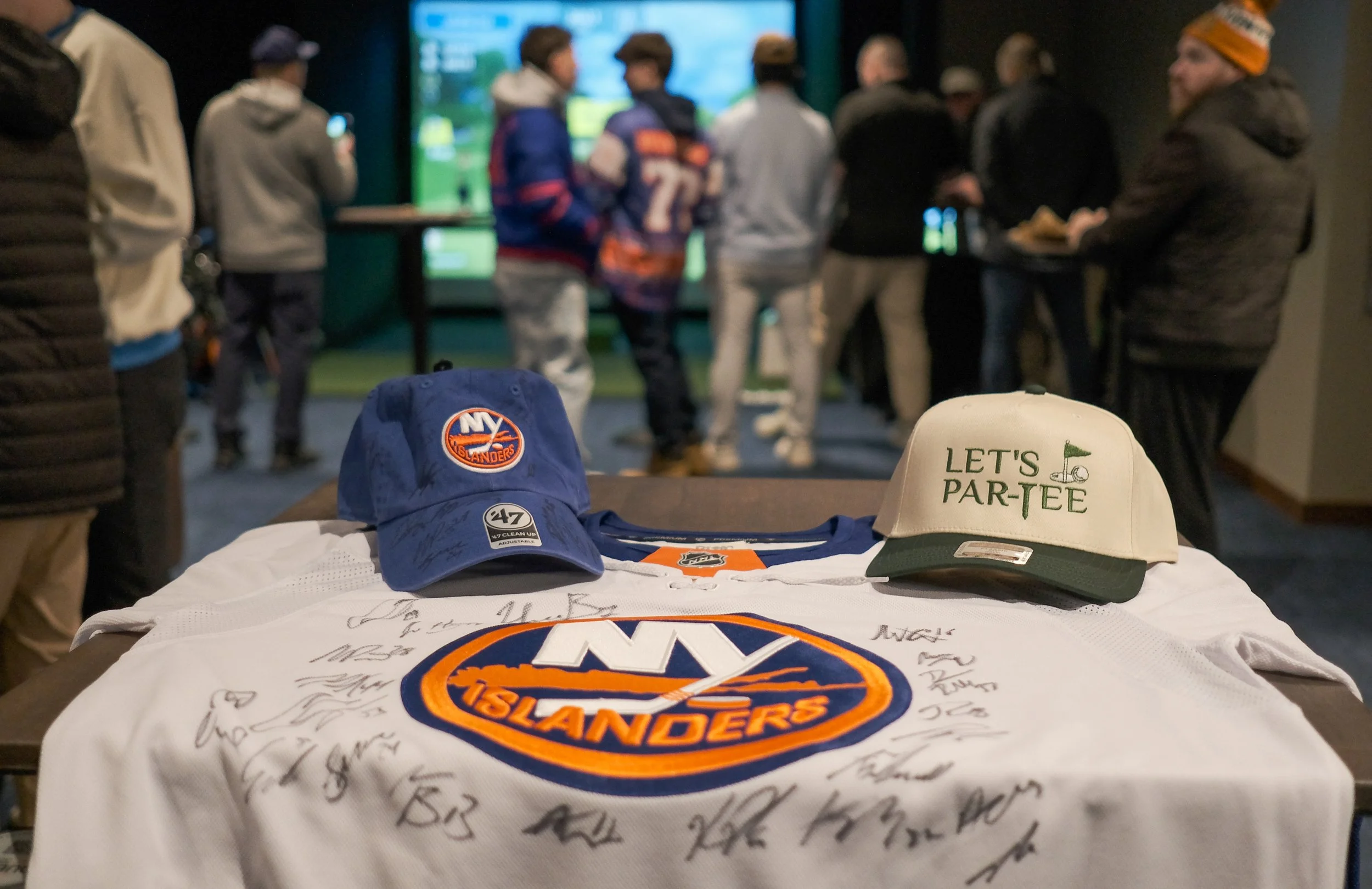 New York Islanders hockey jersey, signed, with two caps on top; one blue with Islanders logo and one white with 'Let's Par-Tee' and a golf flag logo, on a table.