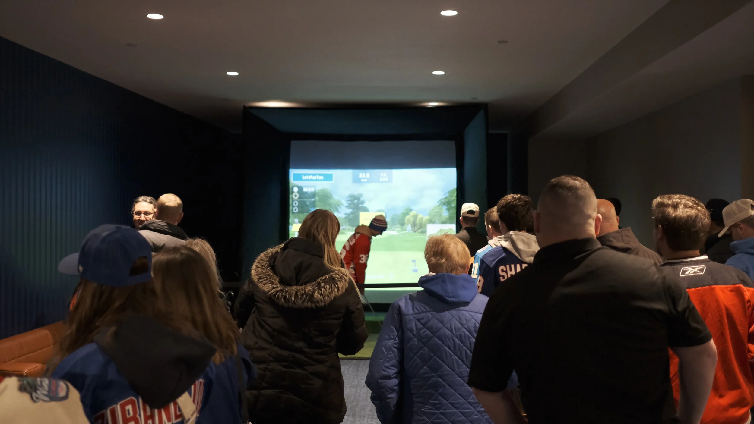 Group of people watching a golf simulation in an indoor room with a large screen.