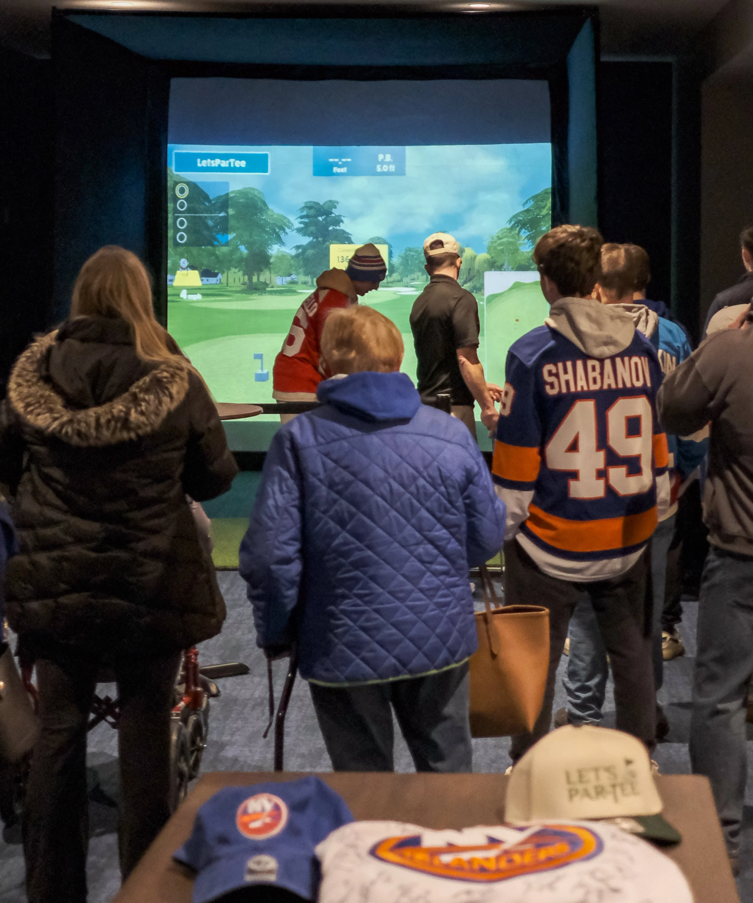 People gathered around a golf simulator screen in an indoor facility.