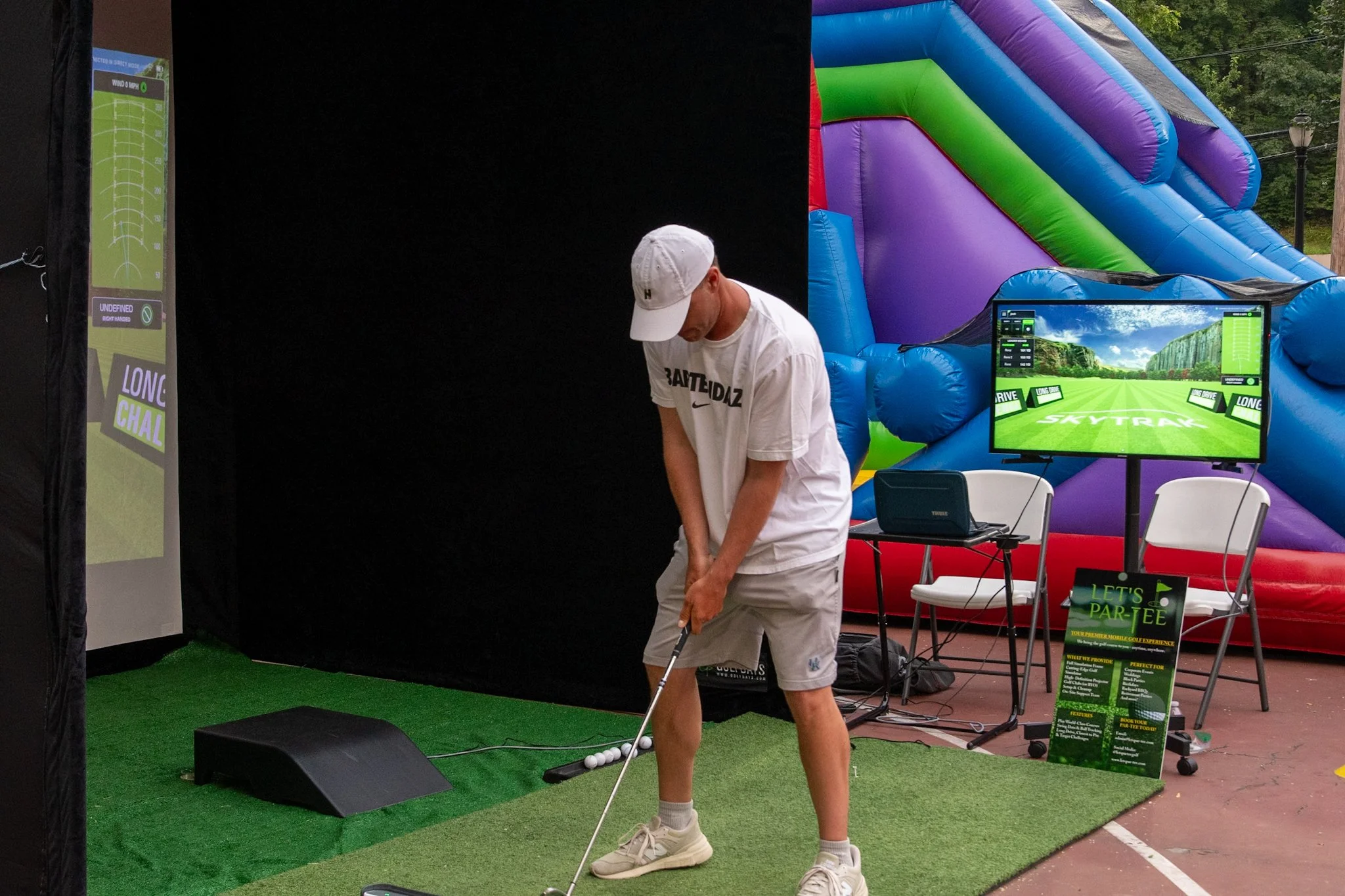 A young man practicing golf swings at an outdoor golf simulation station with an inflatable slide in the background.