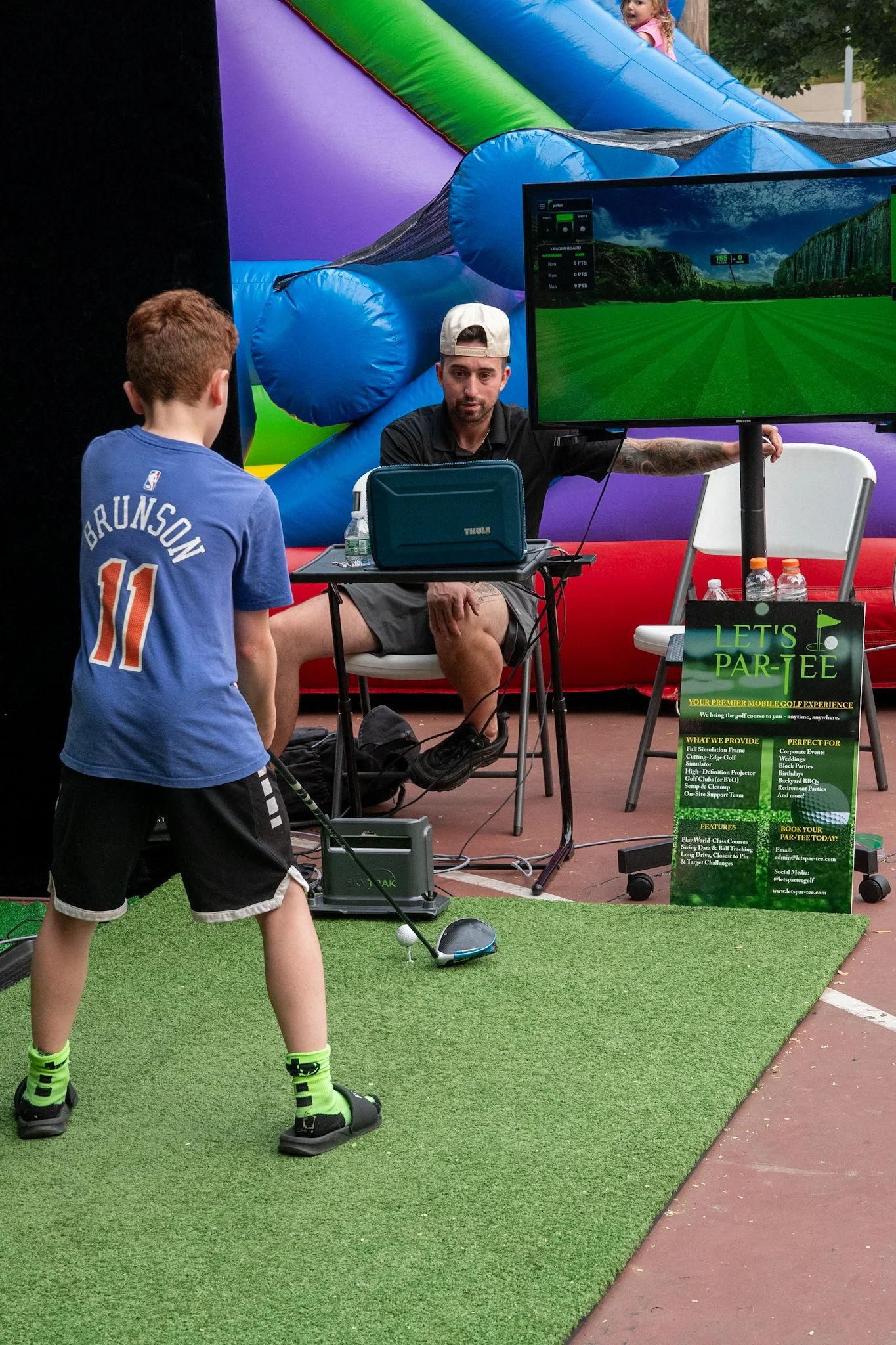 A young boy playing indoor golf on a simulator while an instructor observes, with a large TV screen showing a golf course, and a promotional sign near the golfing area.