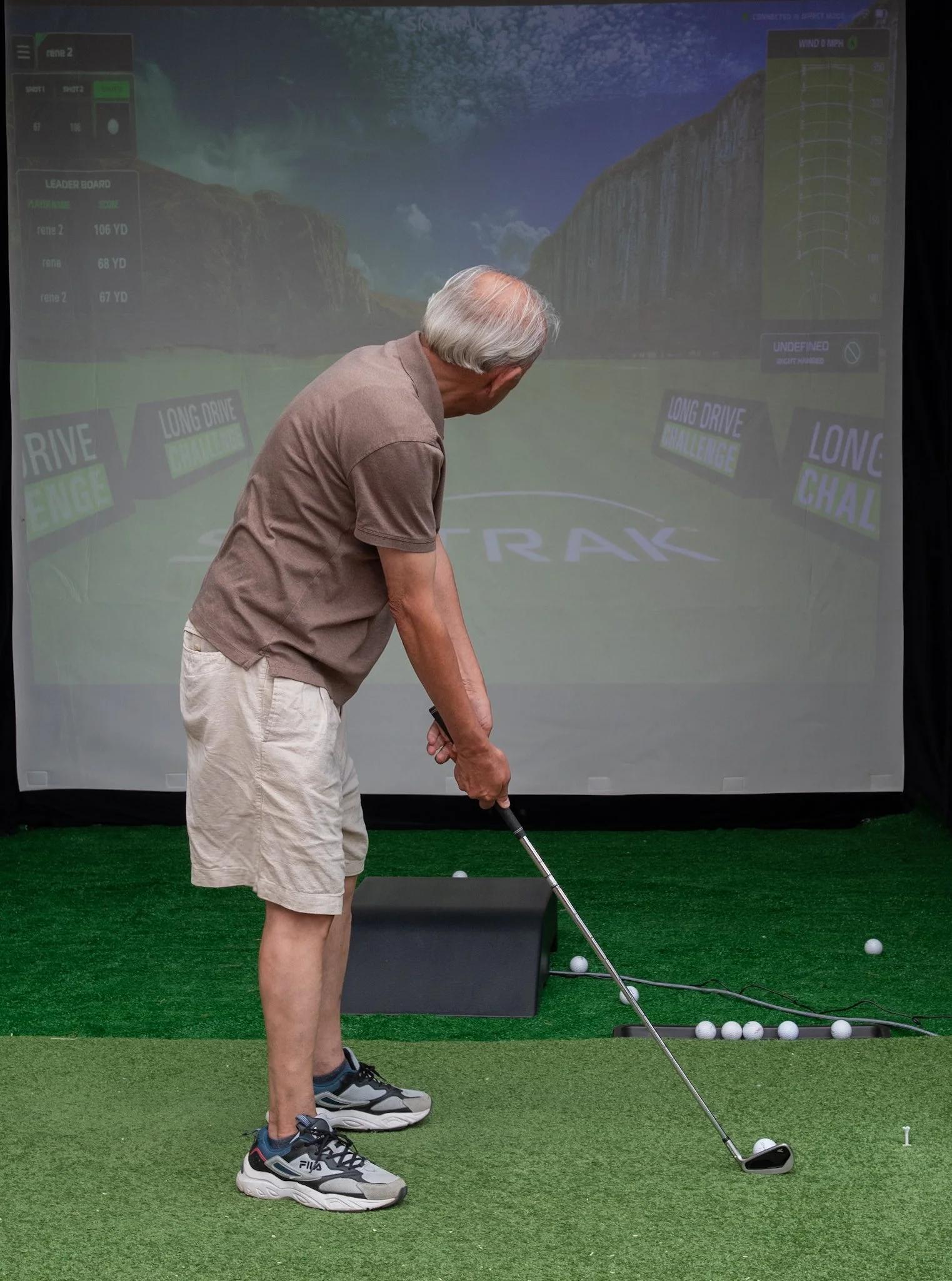 An elderly man practicing golf in a simulated golf environment, standing on artificial turf, preparing to hit a golf ball with a club. There is a large screen in the background displaying a virtual golf course.