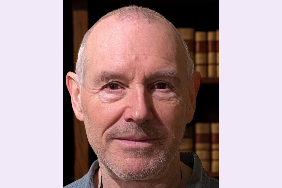 Close-up of a middle-aged man with short gray hair, slight beard, and a neutral expression, in front of a bookshelf filled with books.