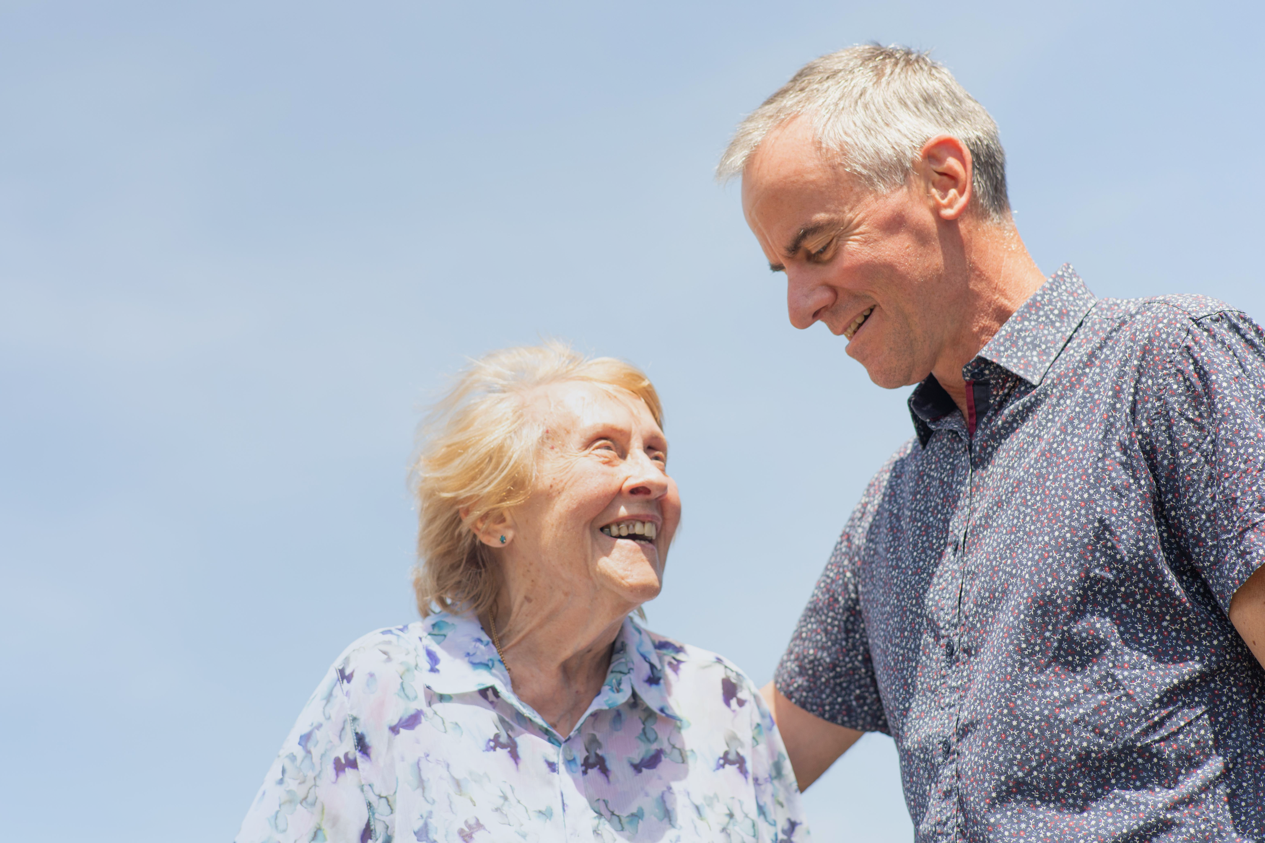 An elderly woman and a middle-aged man are smiling and looking at each other outdoors against a cloudy sky.