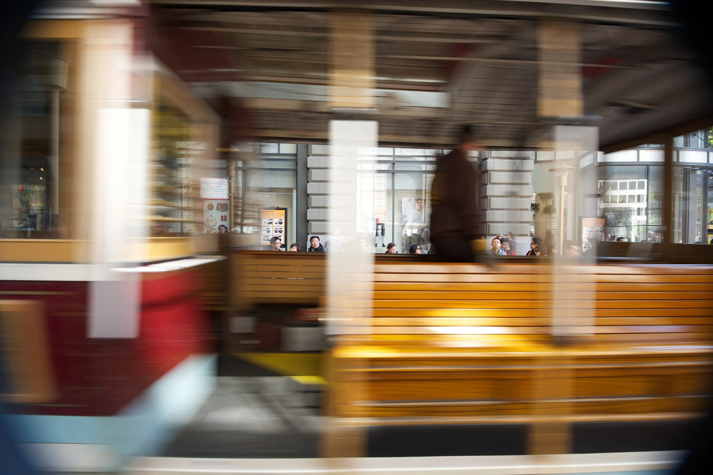 Motion blurred view of a city street with a glass building and people inside, seen through a moving vehicle window.