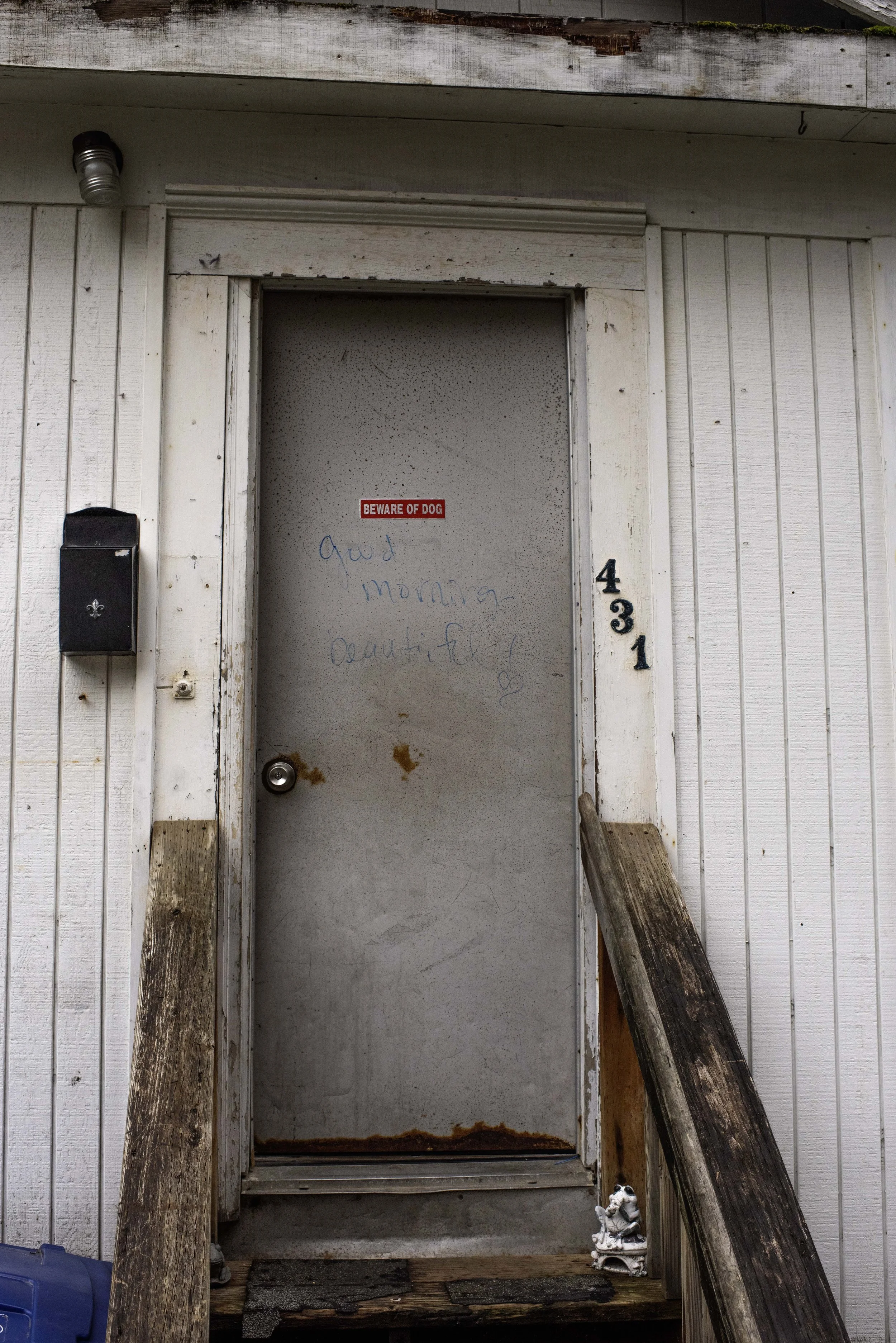 A weathered front door with graffiti, surrounded by white wooden siding, with a small gargoyle statue at the bottom right, and a ramp on the right side.