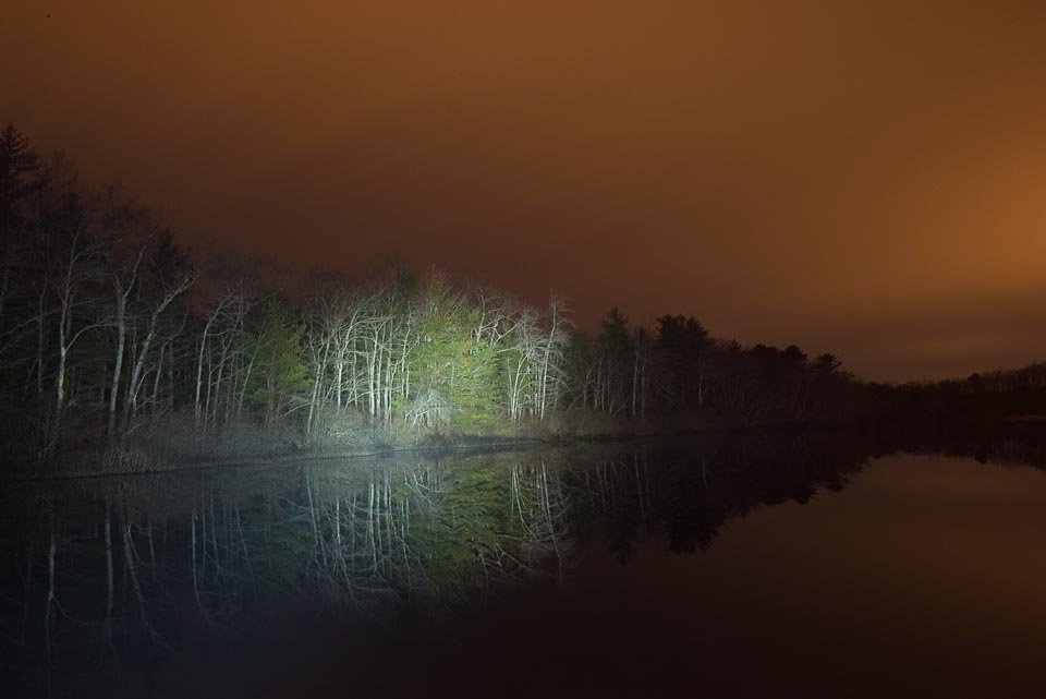 Nighttime scene of a lake with trees on the shoreline illuminated by a bright flashlight, reflecting on the water under a cloudy, orange sky.