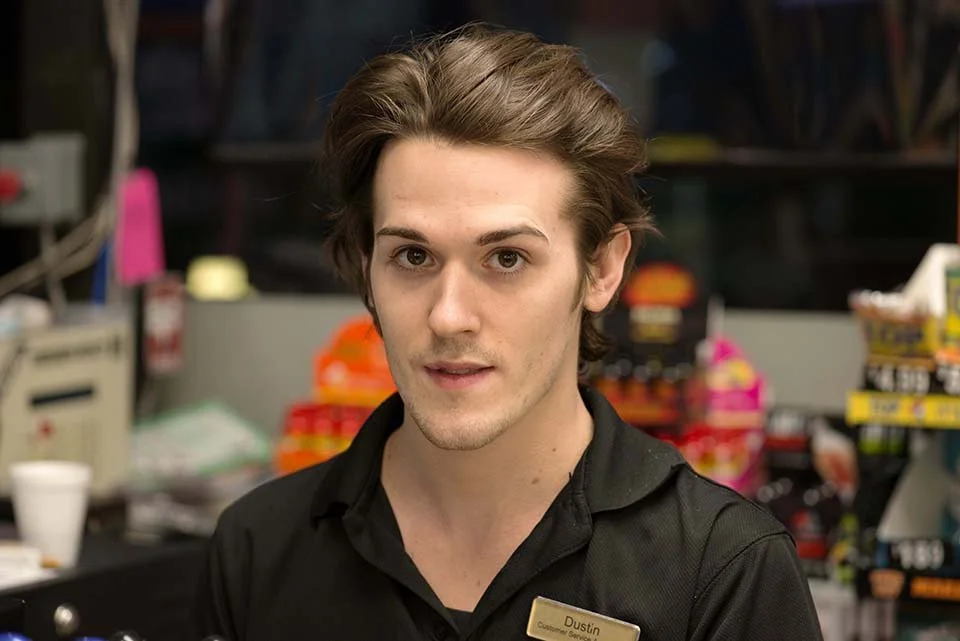 A young man with brown hair wearing a black shirt and a name tag in a retail store.