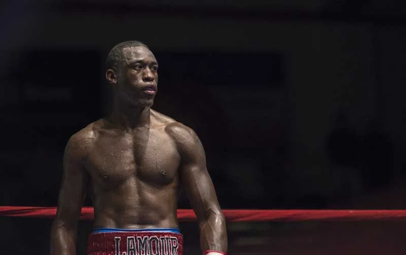 A muscular male boxer standing in a boxing ring, wearing red trunks with blue trim and the word 'L'AMOUR' visible.