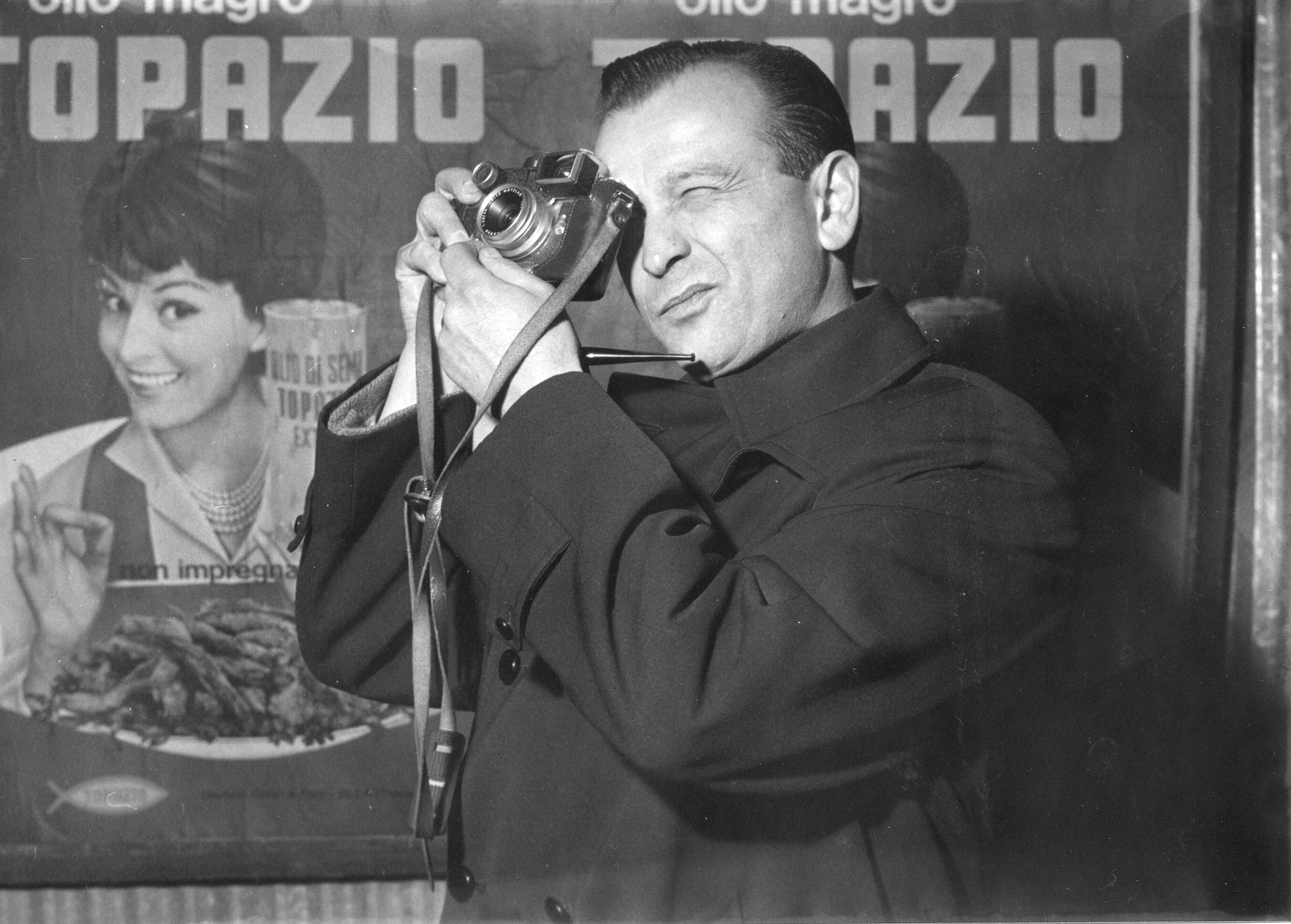 Black and white photo of a man using a camera to take a picture, with a woman smiling in the background, and a table with a large dish of food and a poster with text partially visible behind them.