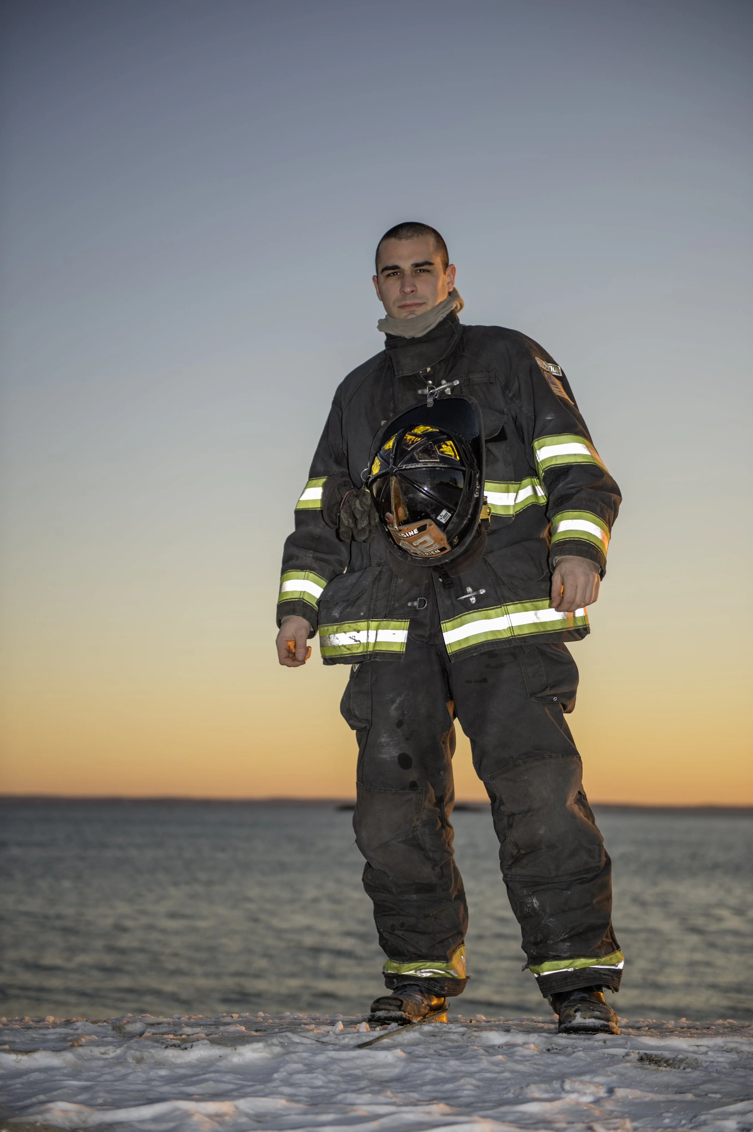 Firefighter standing on snow near water during sunset, holding helmet, looking at camera.