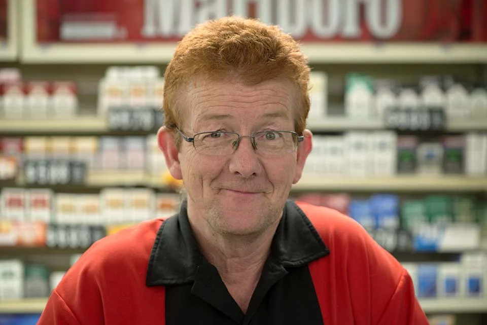 A man with glasses and reddish hair smiling in a store aisle with shelves of medical or pharmacy products in the background.