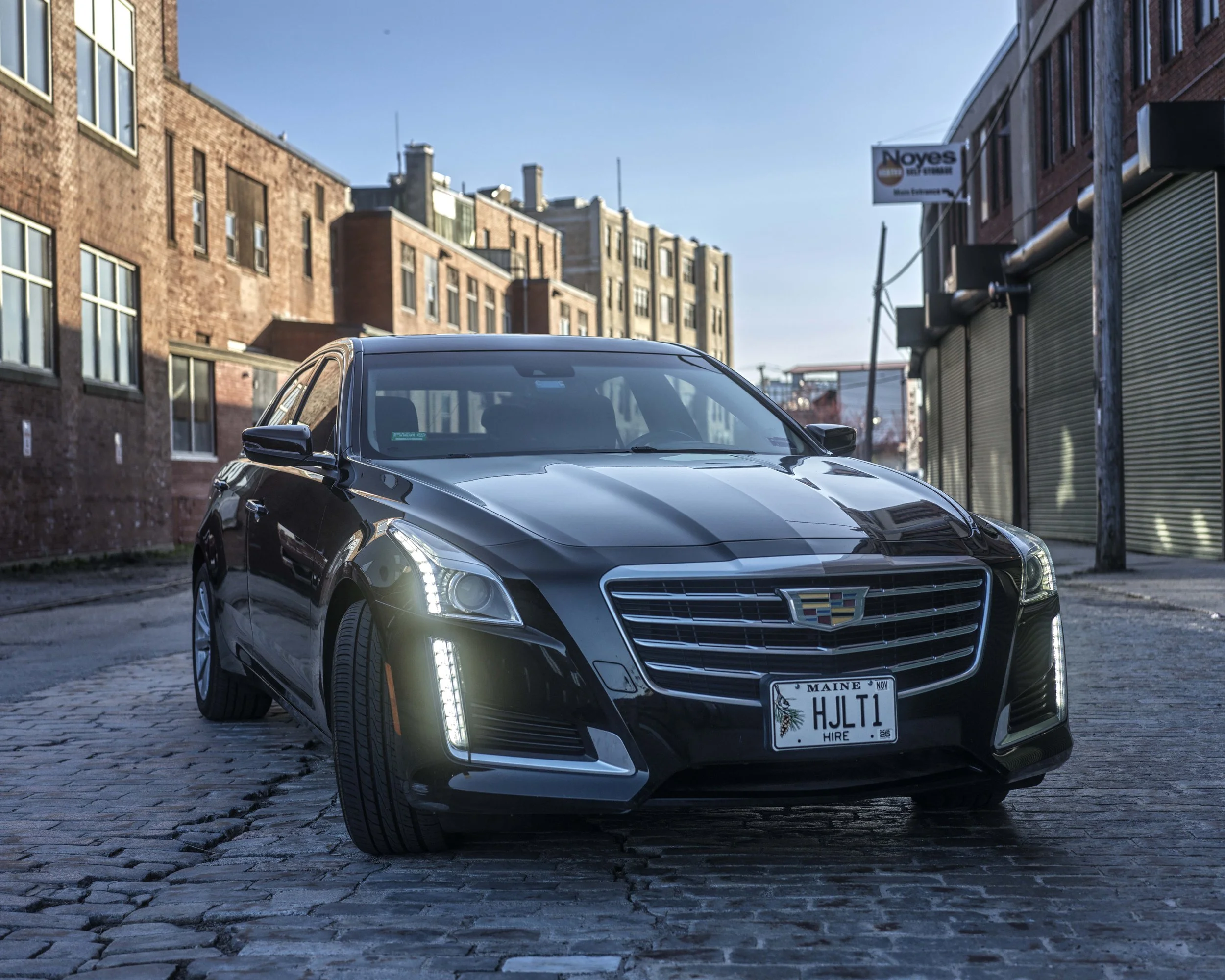 A sleek black Cadillac car with illuminated vertical daytime running lights parked on a cobblestone street in front of brick buildings under a clear blue sky.