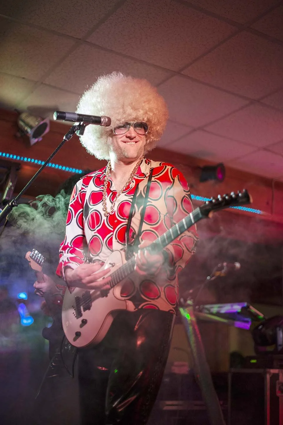A musician with an afro wig, sunglasses, and a colorful 70s-style shirt playing the electric guitar on stage, with a microphone in front of him and fog effects.