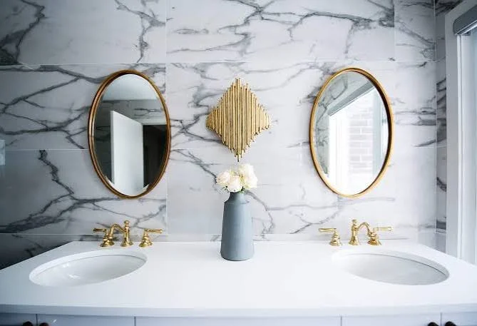 Bathroom vanity with two oval mirrors, a gray vase with white flowers, marble wall background, and two gold-finished sinks and faucets.