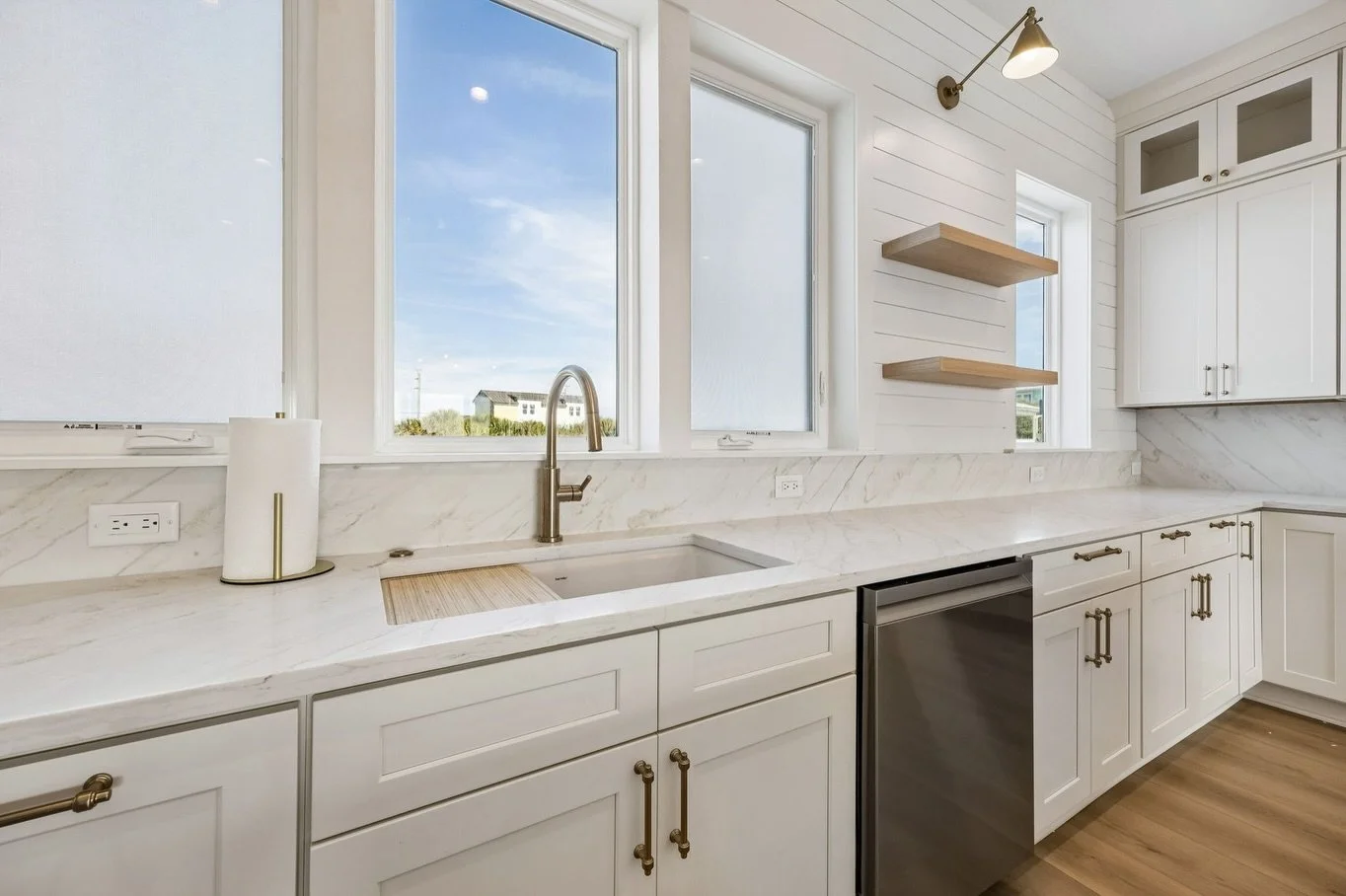 Bright kitchen with white cabinets, marble countertops, and large windows overlooking a cloudy sky and houses outside. Open shelving and a brass wall-mounted light fixture are visible.
