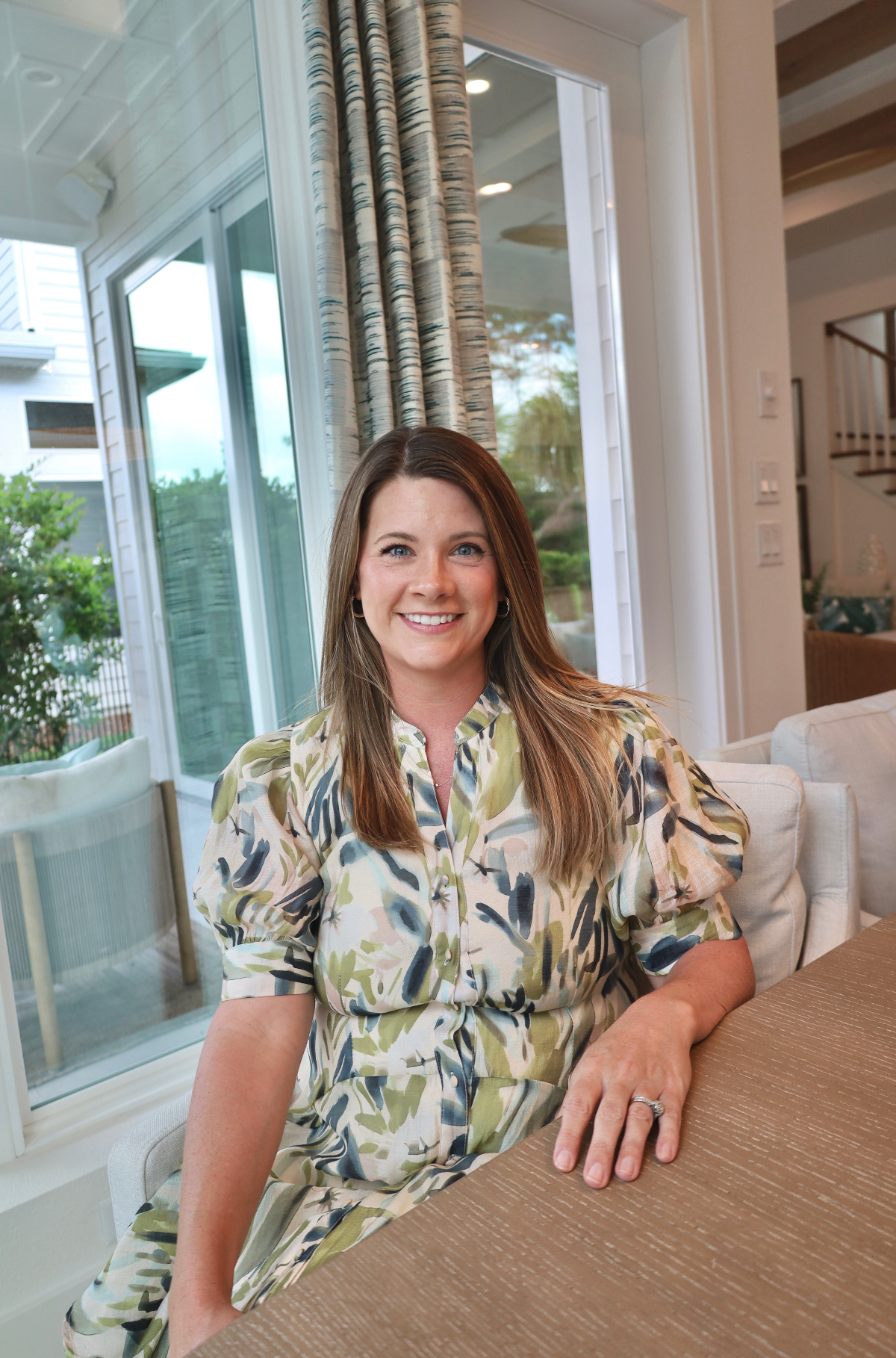 A woman with long, light brown hair and blue eyes sitting at a dining table in a bright, modern room with large windows and curtains. She is smiling, wearing a patterned short-sleeve dress.