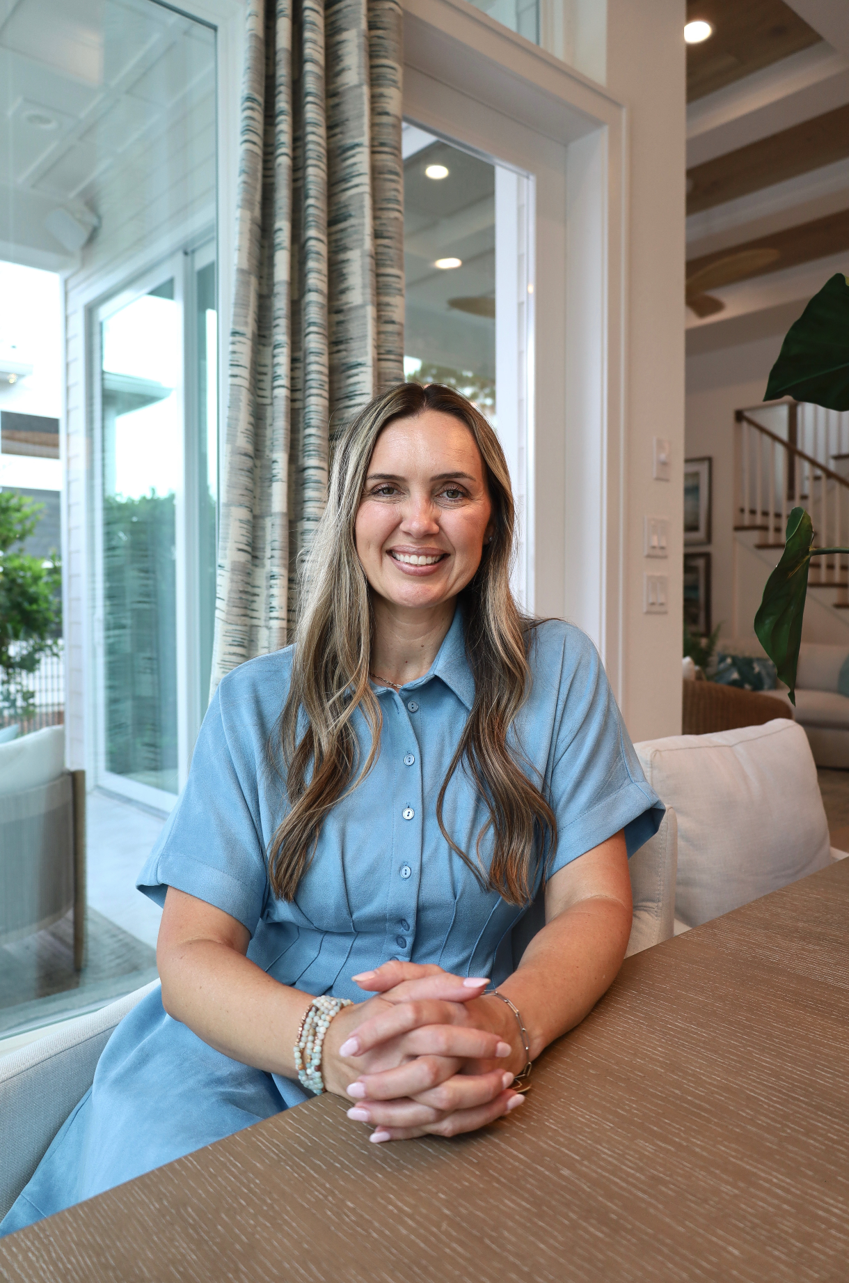 A woman with long wavy light brown hair and light skin is smiling and sitting at a dining table with her hands clasped in front of her. She is wearing a light blue button-up shirt and beaded bracelets on her left wrist. Behind her, there is a large window with patterned curtains, a view of the patio, and a modern, well-lit interior with stairs and framed pictures on the wall.