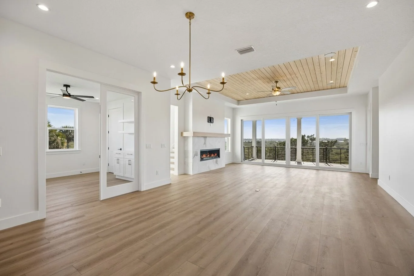 Open living room with wood floors, a built-in fireplace, a chandelier, ceiling fan, large windows and sliding door to balcony, and a sitting area with a wooden ceiling.