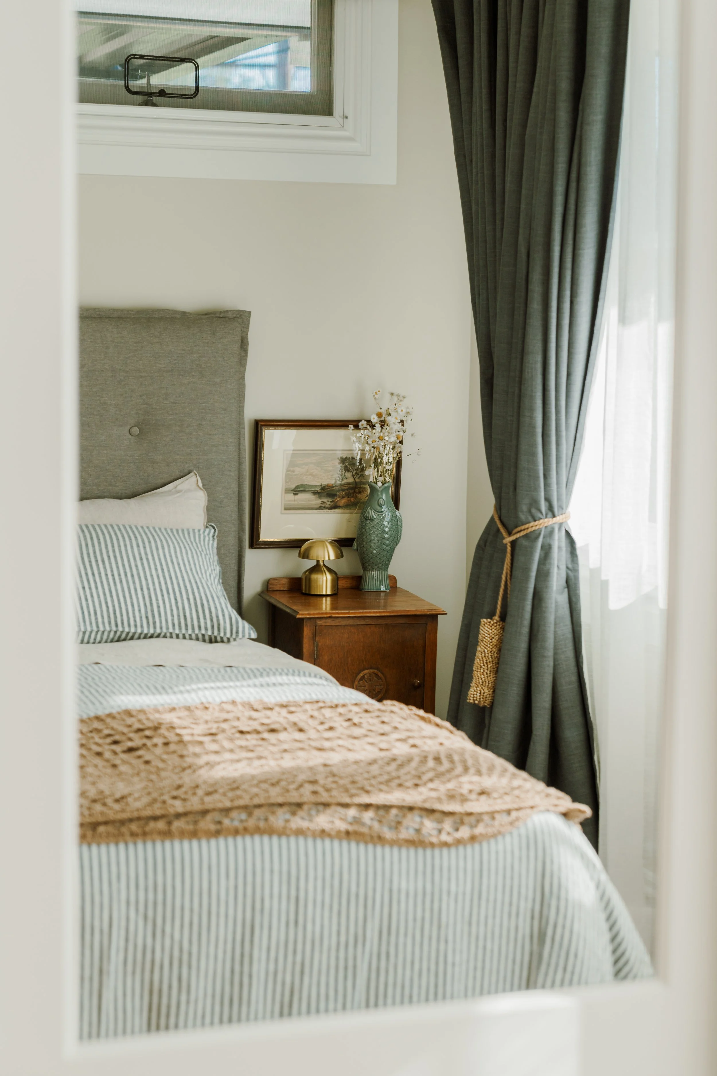 The reflection of a well made bed, bedside table with a vase and small light and curtain