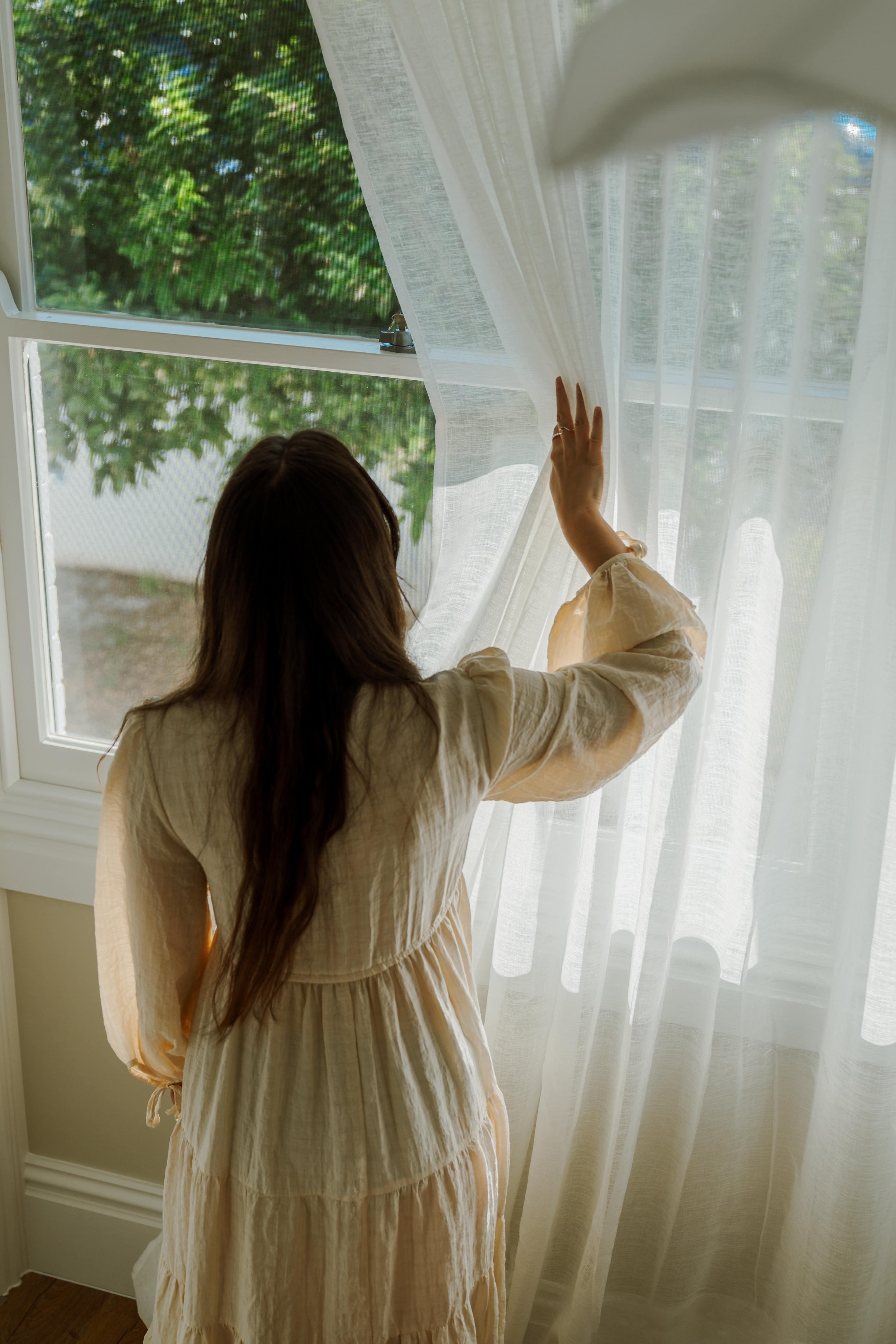 Girl in linen dress holds curtain aside and looks out window at a tree