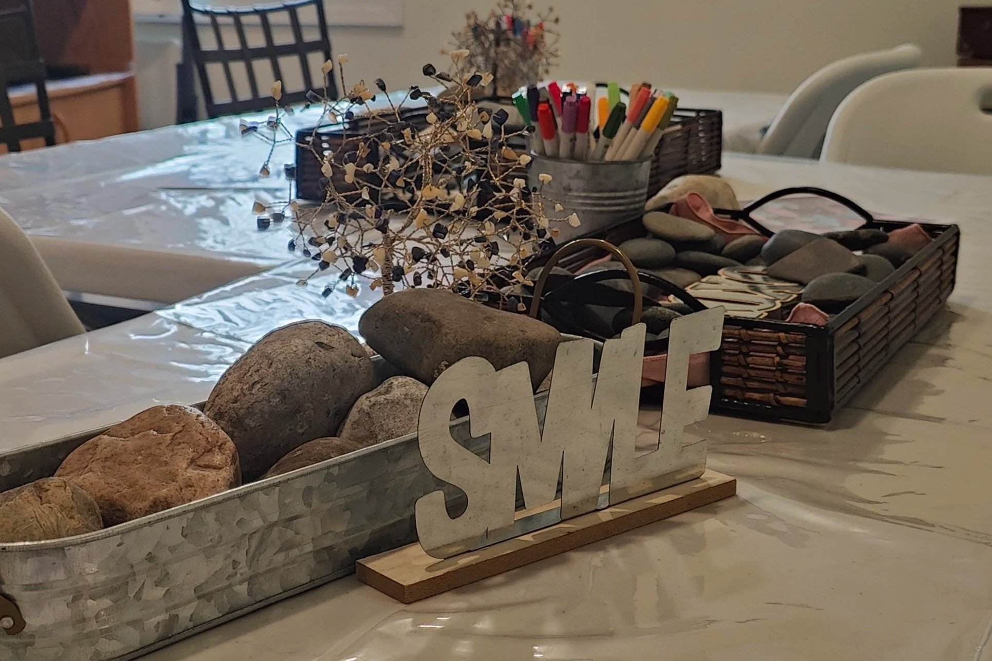 Decorative table centerpiece with rocks, dried plant, and 'SMILE' sign on a dining table with pens and stones in baskets.