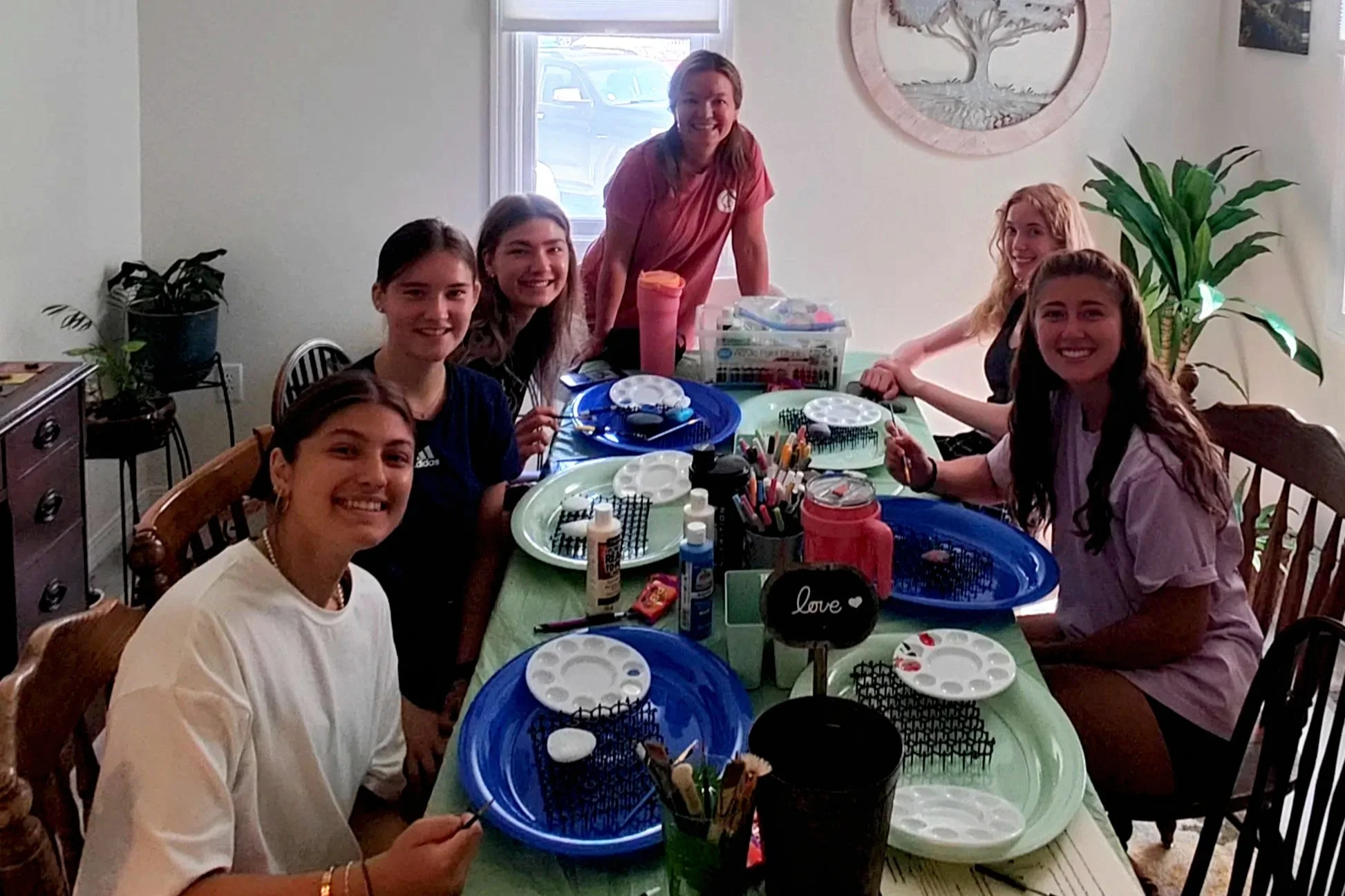 Six young girls sitting around a brightly decorated table, engaged in arts and crafts, with supplies including paint, glue, and paper.