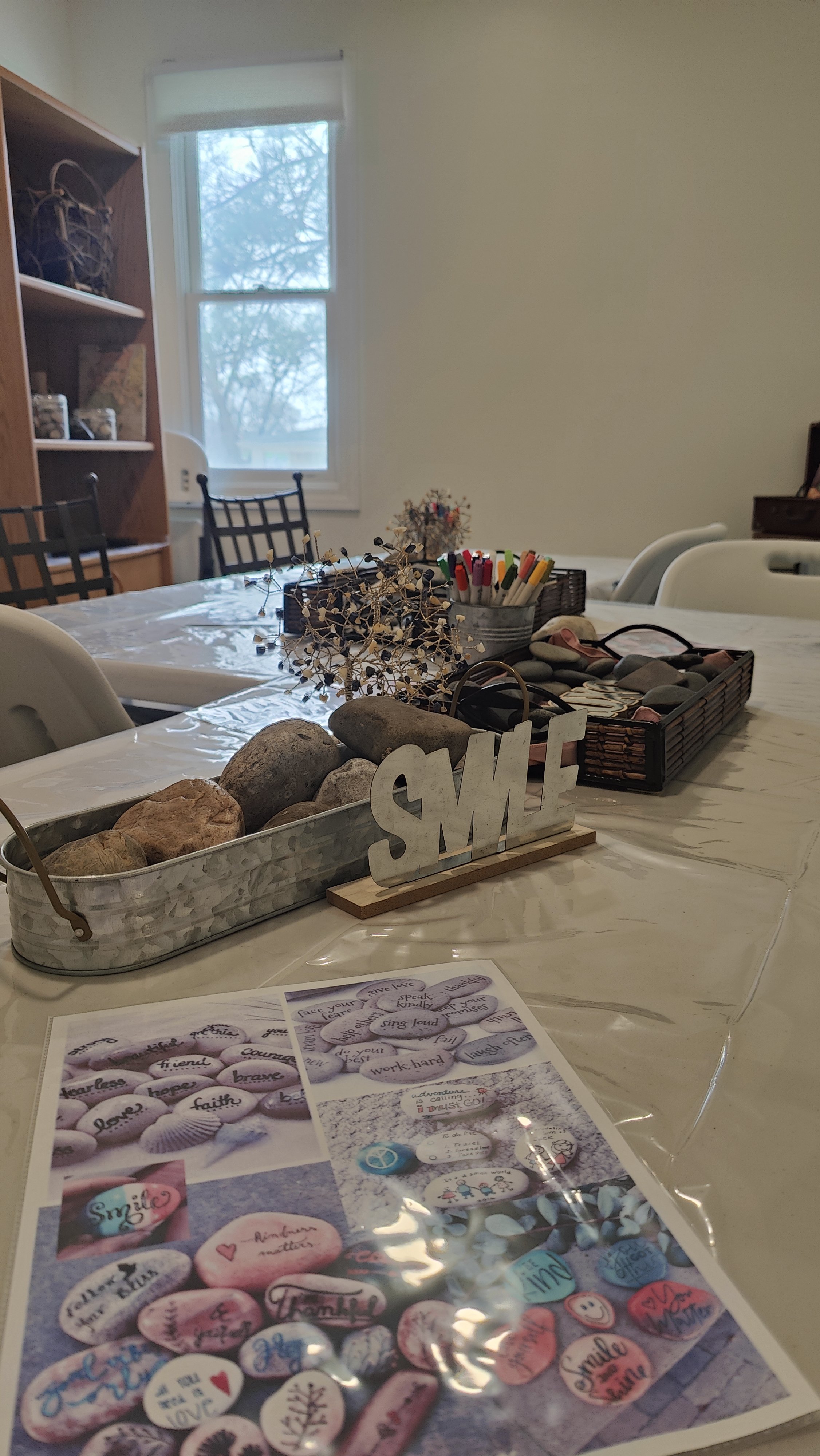 A dining table decorated with a metal tray holding rocks, a "SMILE" sign, and a tray with pens and stationery. The table also has a pamphlet with painted rocks with inspirational words, set in a room with a window, chairs, and a bookshelf.