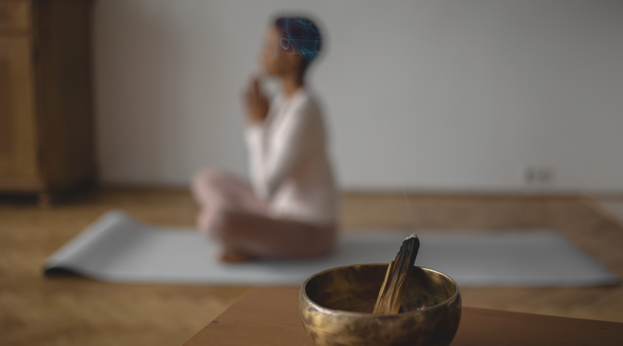 A smoking incense stick in a brass bowl with a woman practicing yoga in the background, sitting cross-legged on a yoga mat.