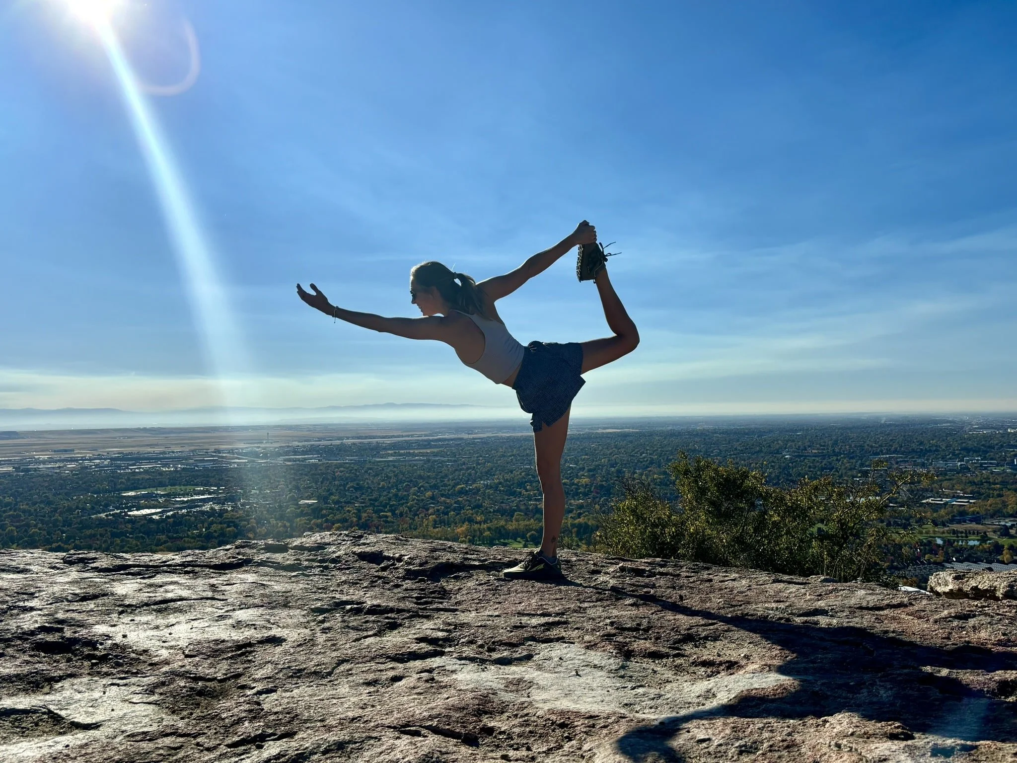 A person doing a yoga pose on a rocky mountaintop with a city and body of water in the distance under a blue sky with sunshine.