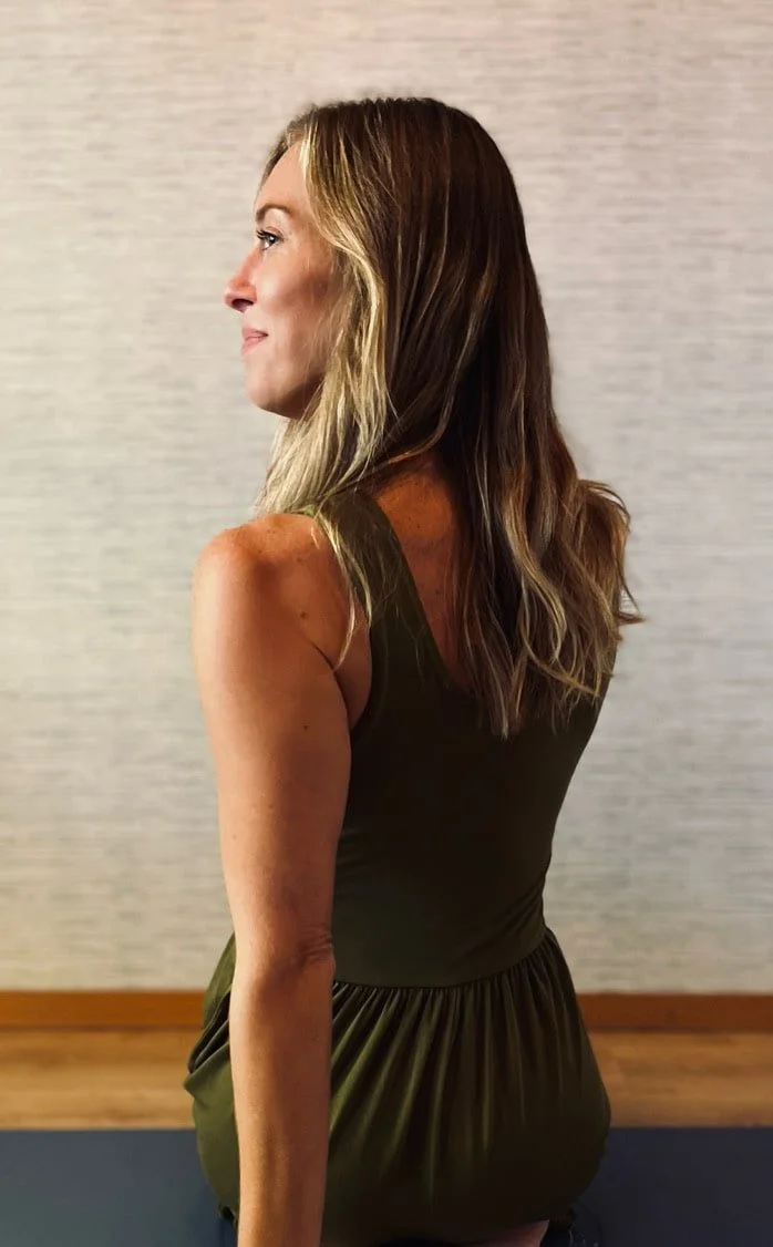 A woman with long, wavy blonde hair, sitting cross-legged on a yoga mat indoors, facing sideways with a calm expression.
