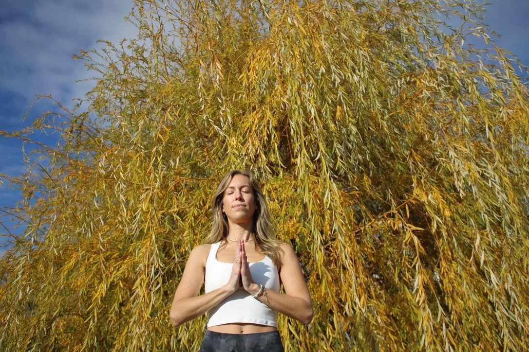 A woman practicing yoga outdoors with a large yellow tree behind her, her hands in a prayer position, eyes closed, in a calm and peaceful setting.