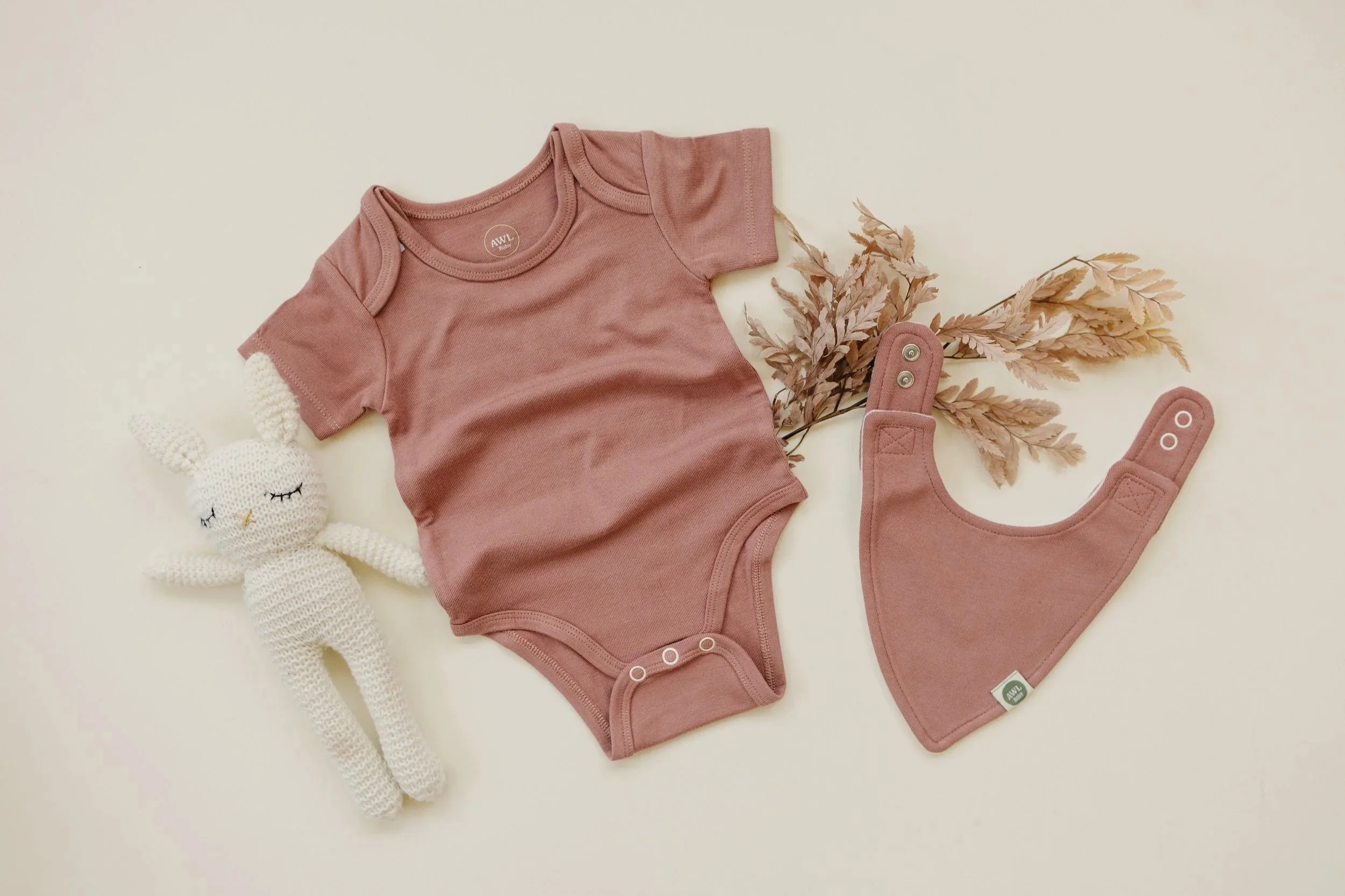 Flat lay of a baby bodysuit with matching quick-change bib in a soft mauve color, styled with a crochet bunny and dried florals.
