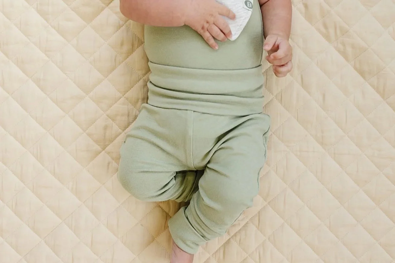Baby wearing a sage green grow-with-me style outfit, lying on a quilted mat.