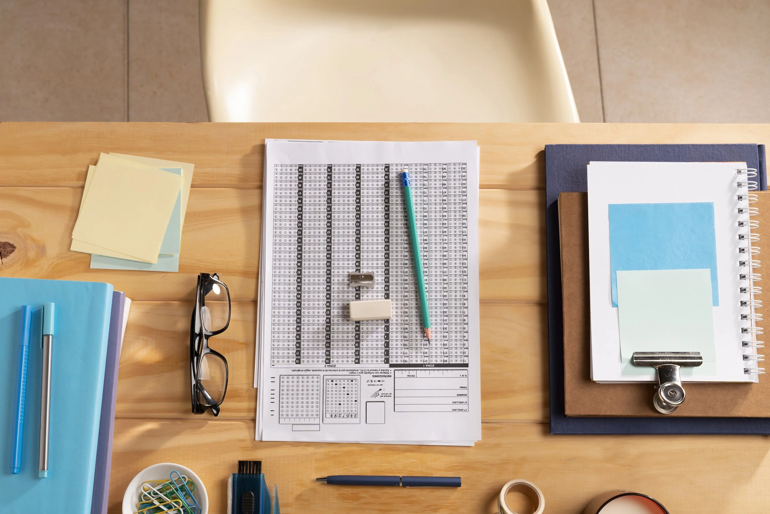 Desk with office supplies including a pair of glasses, sticky notes, a blue folder with pens, a newspaper, a black pen, a small bowl of paper clips, a yellow ink stamp, and notebooks and folders arranged neatly.