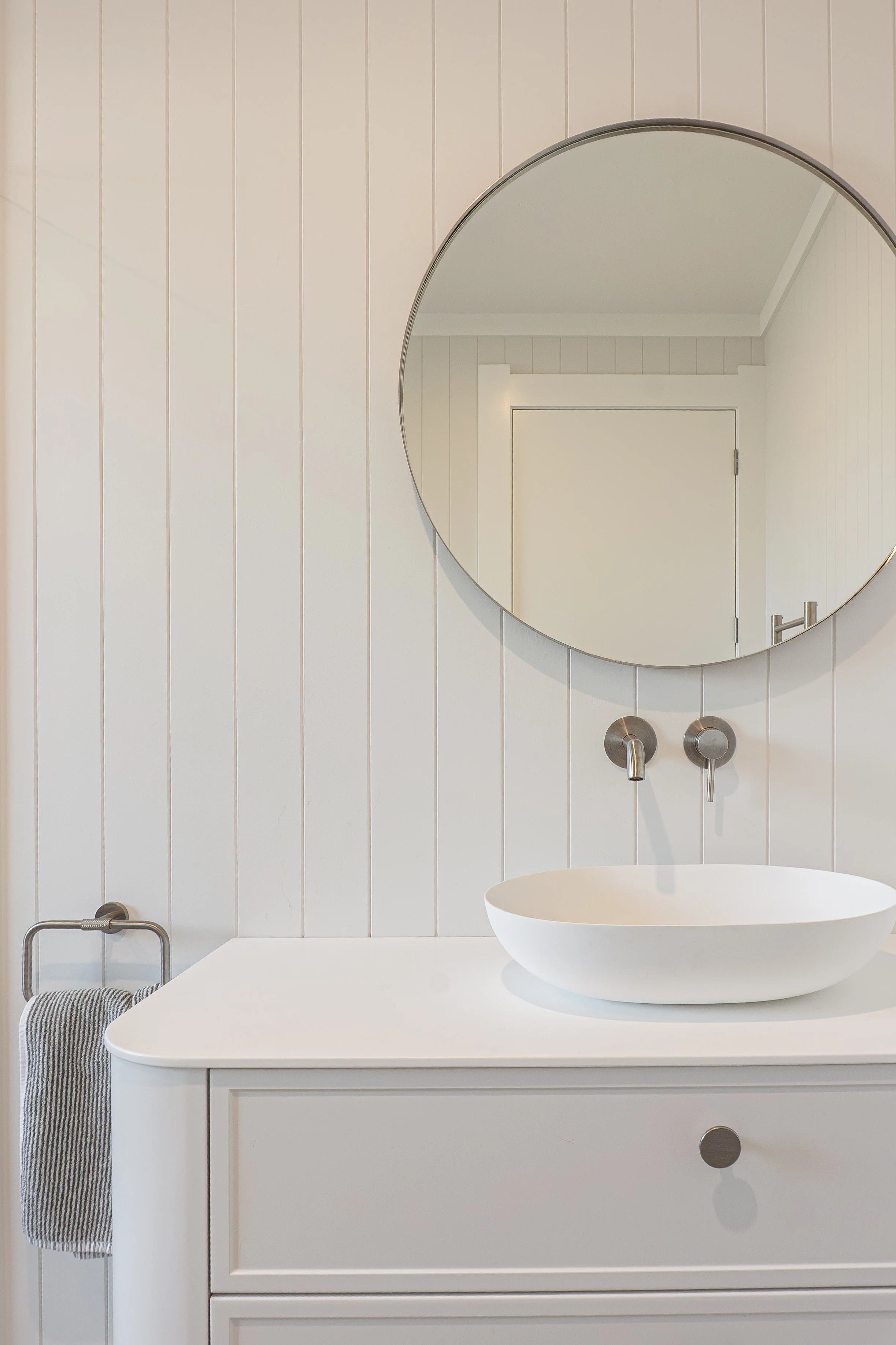 A modern white bathroom vanity with a vessel sink, round mirror, and wall-mounted faucet, with a striped hand towel hanging on a metal towel holder.
