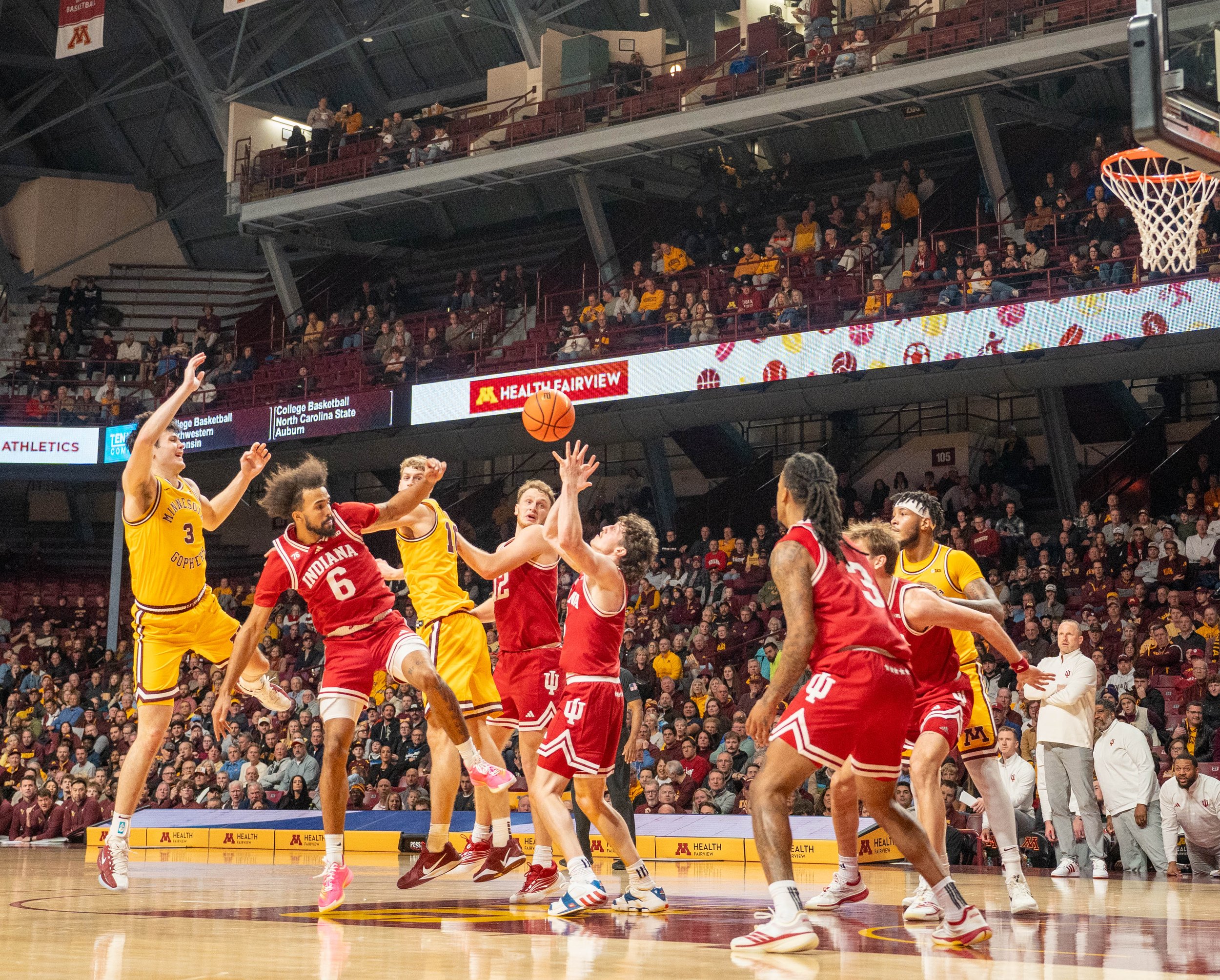 Basketball game in progress with players from Minnesota and Indiana teams on the court, crowd seated in the stands, basketball hoop on the right, and digital advertisements on the wall.