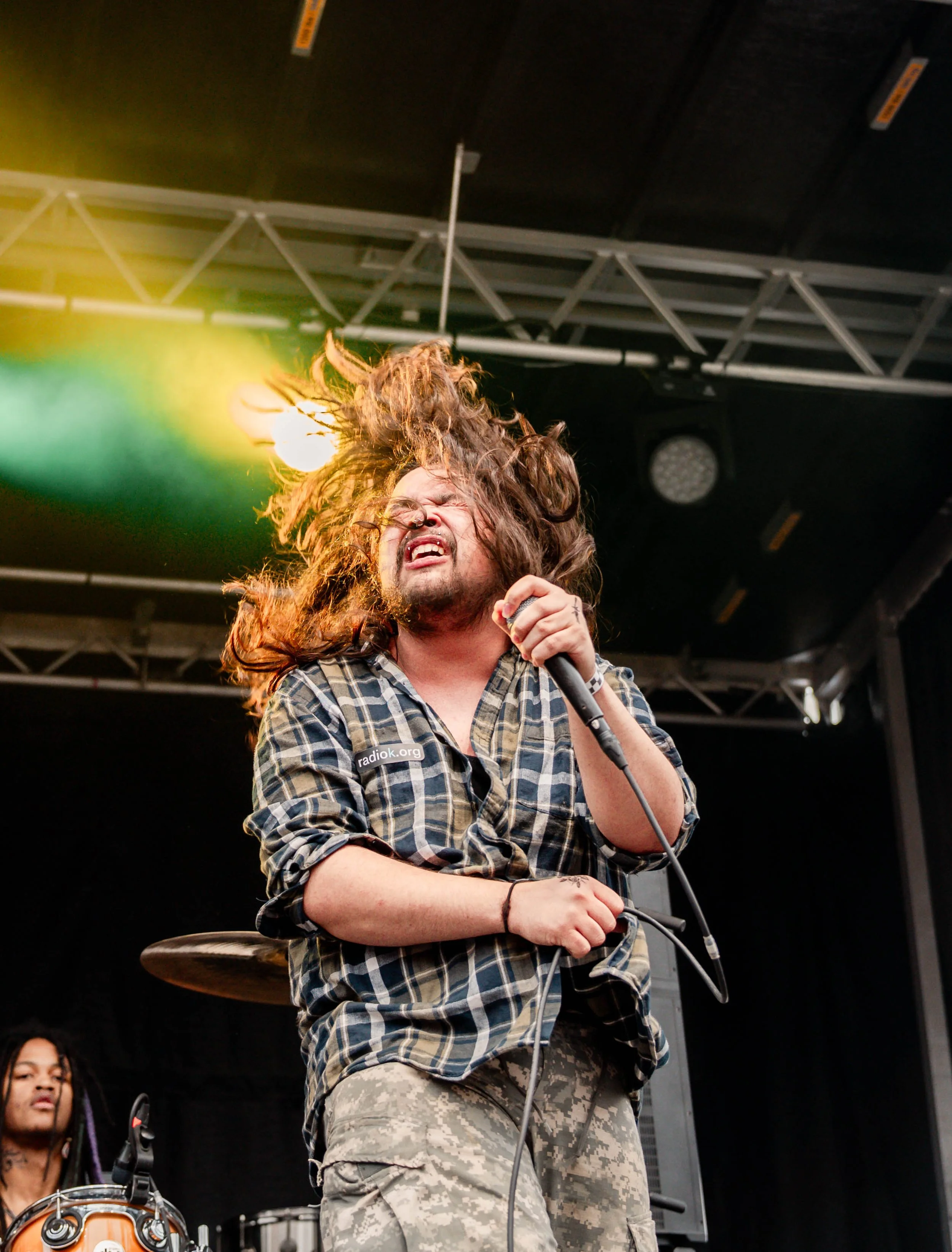 A musician with long curly hair, wearing a plaid shirt and camouflage pants, singing into a microphone on stage with colorful lighting and drums visible in the background.