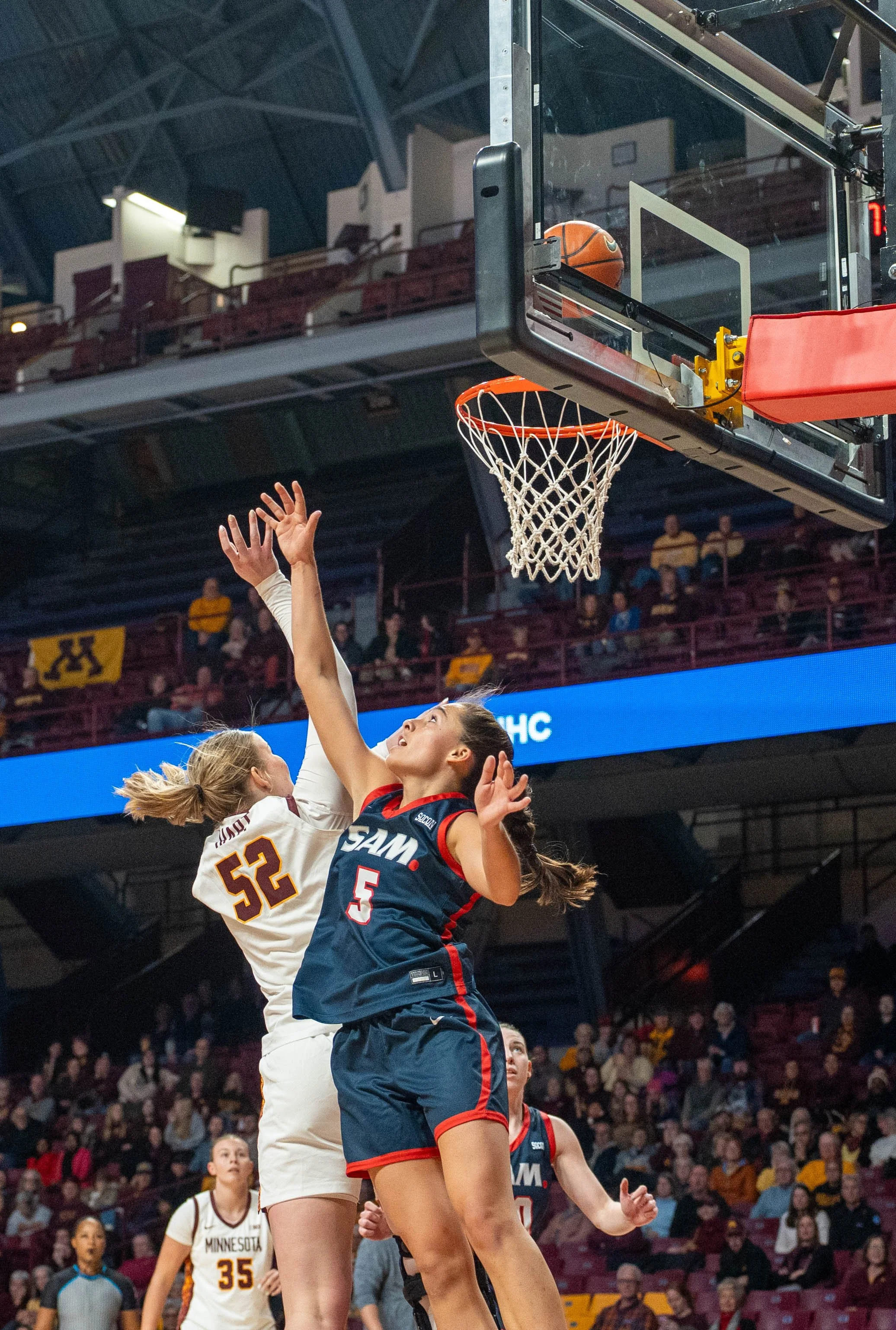 Two female basketball players are jumping to grab the ball near the basketball hoop during a game. Spectators are visible in the background.