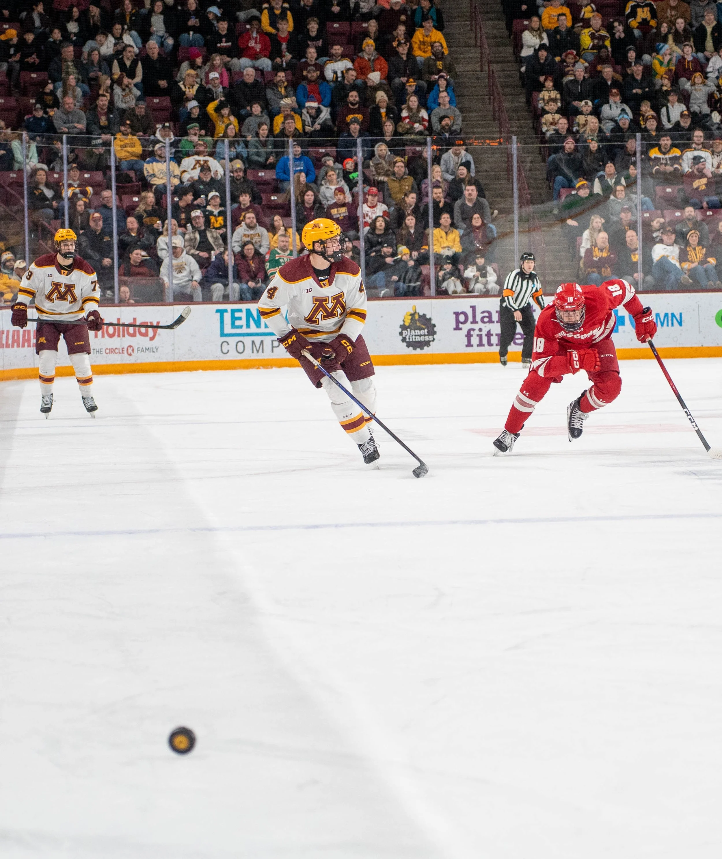 Ice hockey game with players in maroon, white, and red jerseys, spectators in the stands, and a referee on the ice.