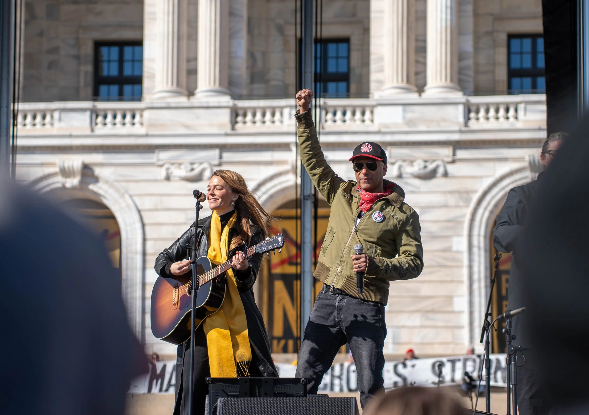 A woman with long reddish hair playing an acoustic guitar and singing into a microphone, with a man in sunglasses and a cap raising his fist, performing on an outdoor stage in front of a large historic building.