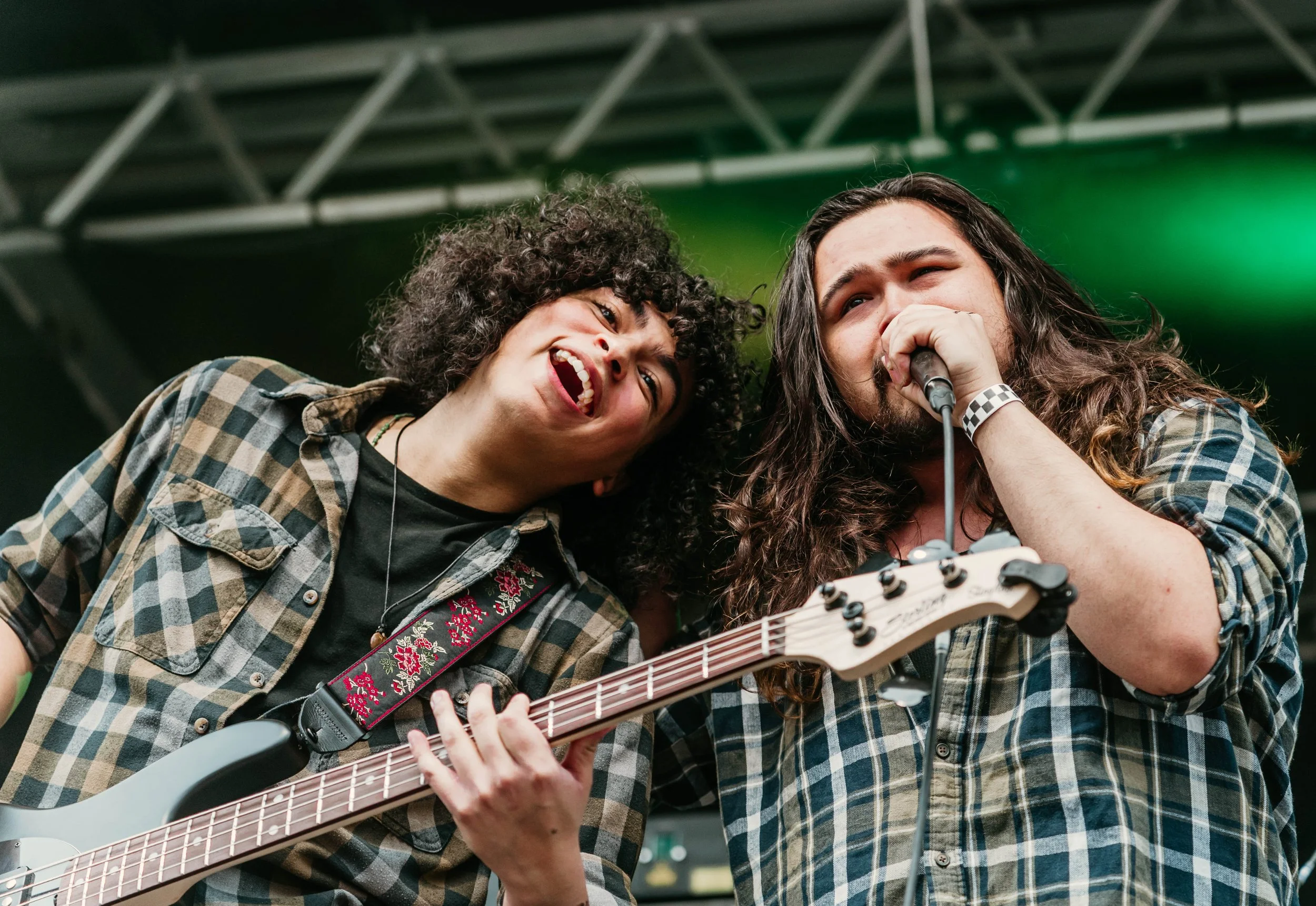 Two musicians performing on stage, one singing into a microphone and the other playing an electric guitar, both wearing plaid shirts.