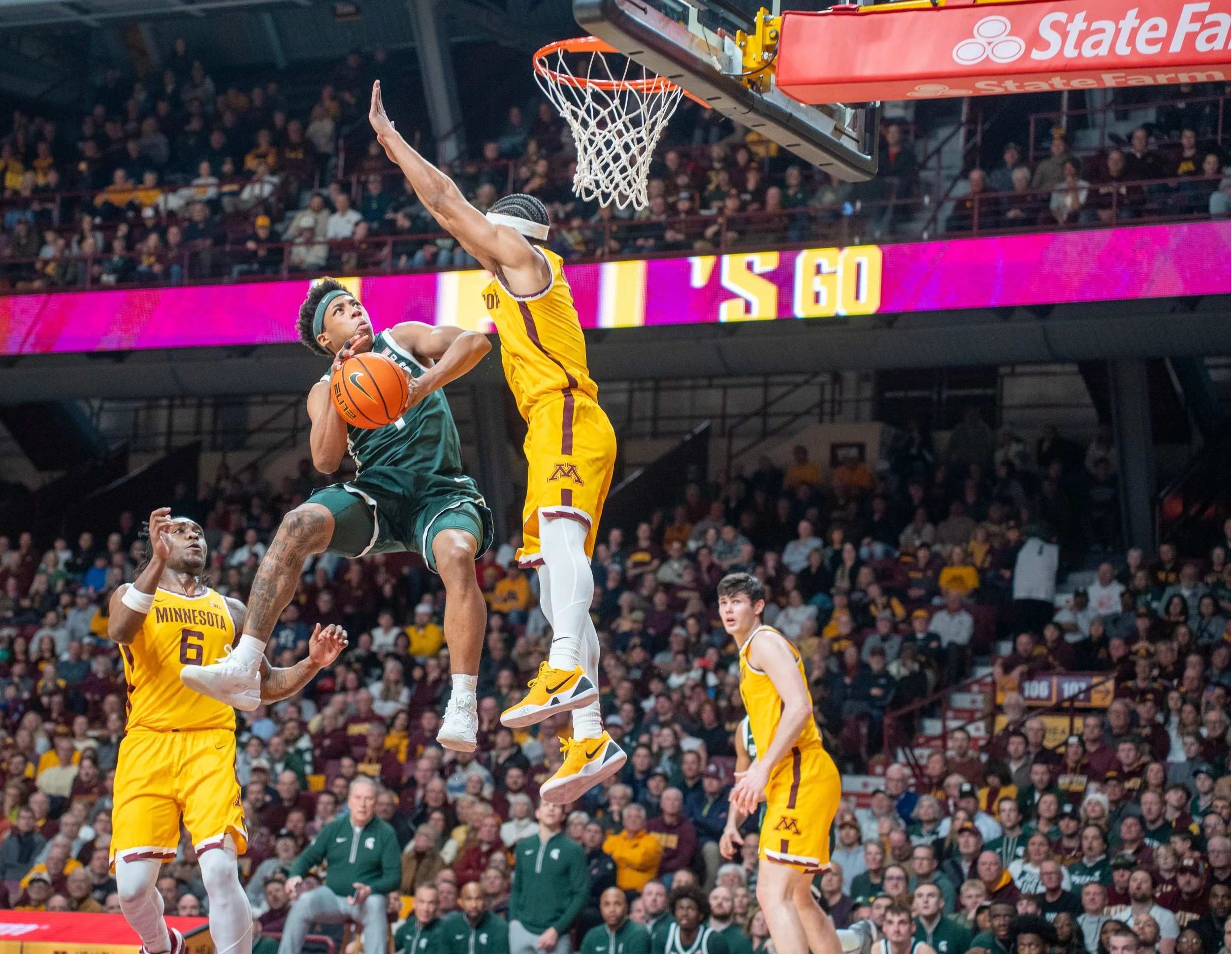 A college basketball game between teams from Michigan and Michigan State. Two players are jumping for a rebound near the basket, with a crowd of spectators watching in the background.