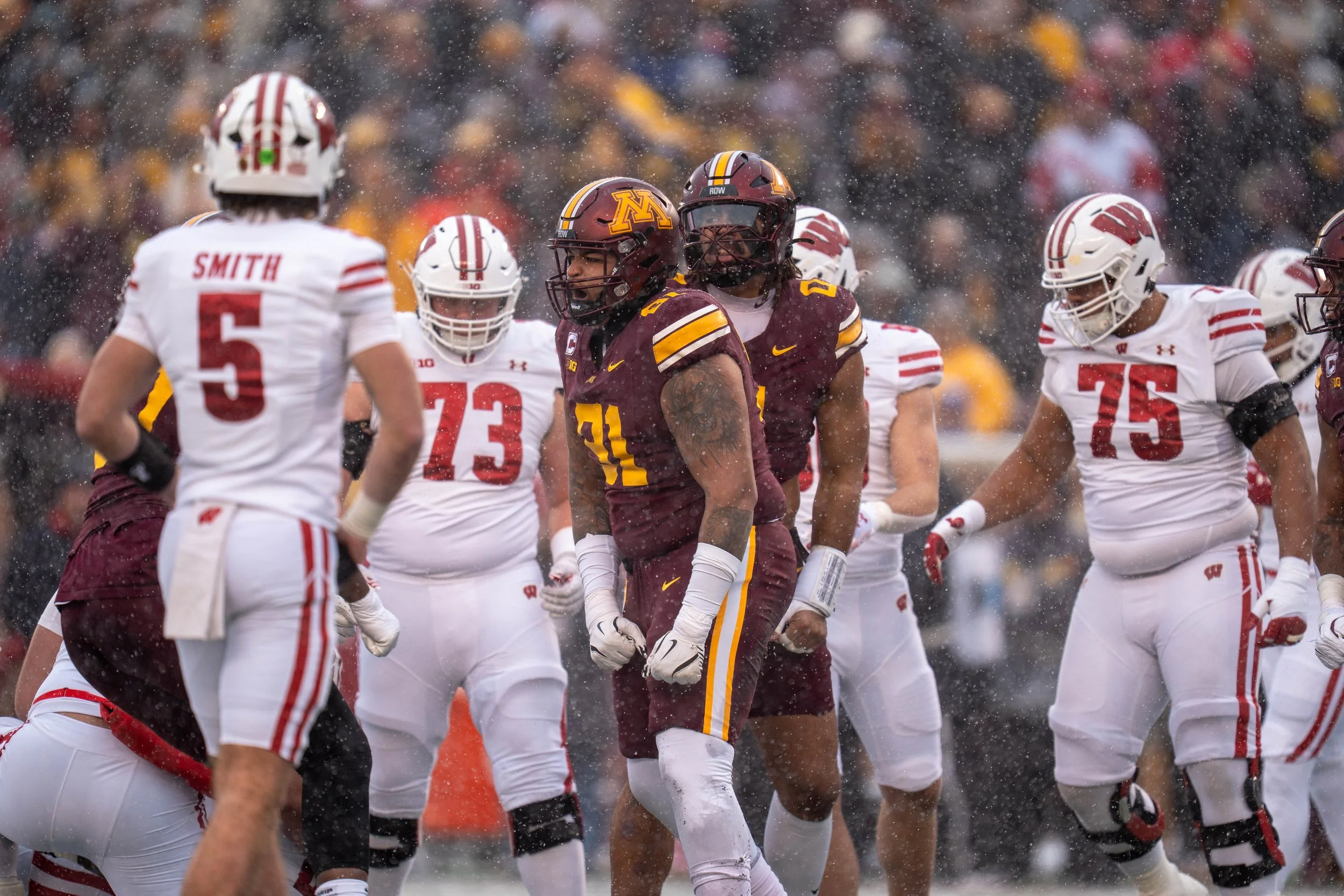 Group of American football players, some wearing white and red uniforms and others wearing maroon and gold, standing and talking on the field during a rainy game.
