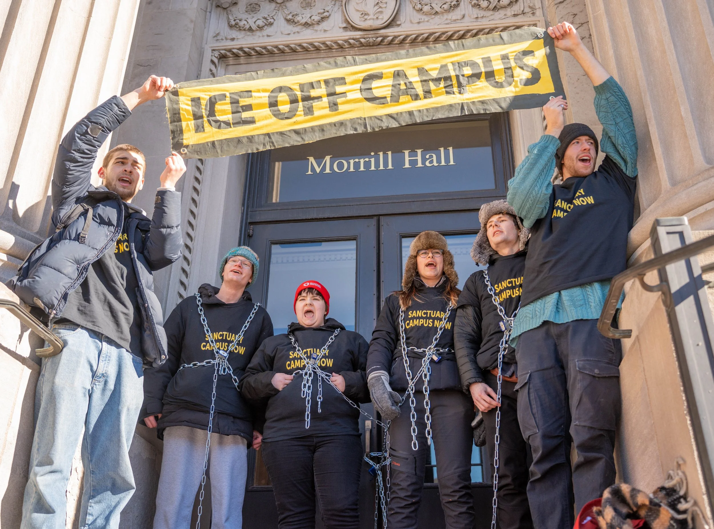 Protesters standing on stairs outside Morrill Hall holding a 'ICE OFF CAMPUS' banner, with some wearing chains and matching black shirts that read 'Sanctuary Campus Now'.