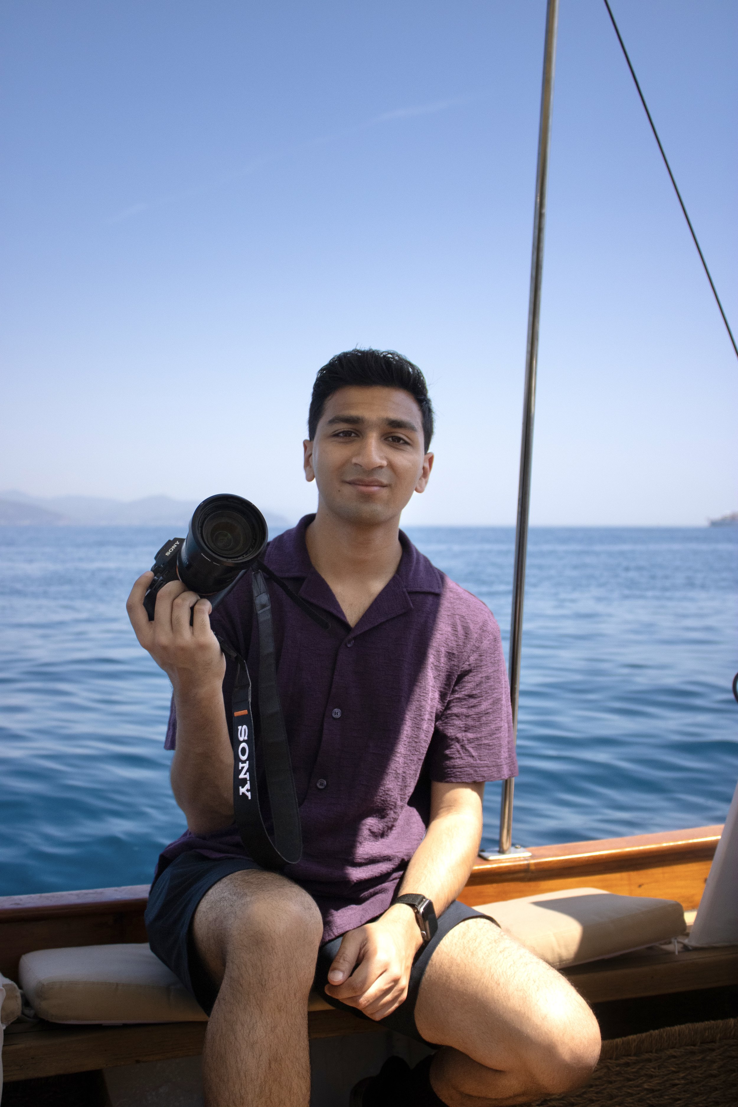 A young man sitting on a wooden bench on a boat, holding a camera in his right hand, with water and distant hills in the background, under a clear blue sky.