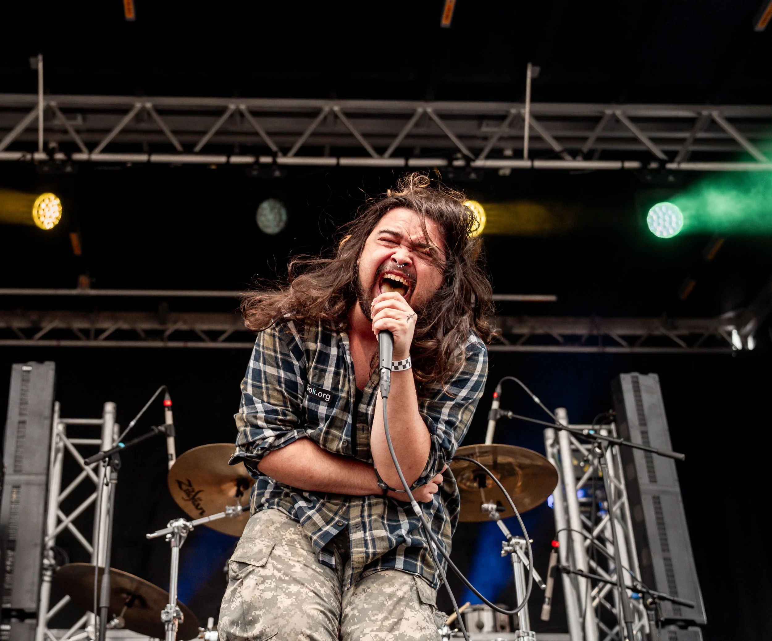 Male singer with long hair and beard singing passionately into a microphone on stage, with drum set and stage lights in the background.