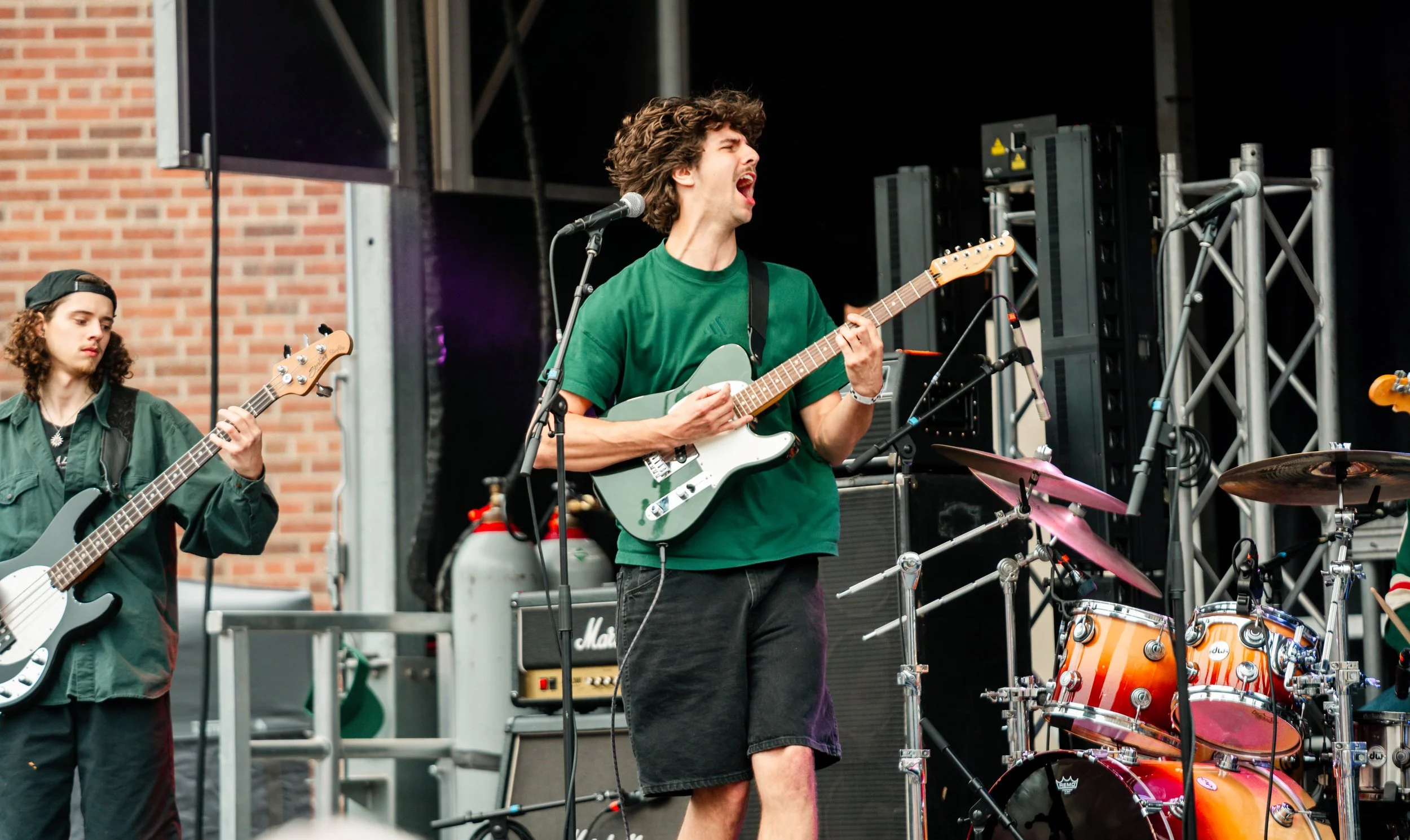 Musicians performing on an outdoor stage, one with curly hair playing an electric guitar and singing, another with long hair and a cap playing a bass guitar, with musical equipment and brick wall background.